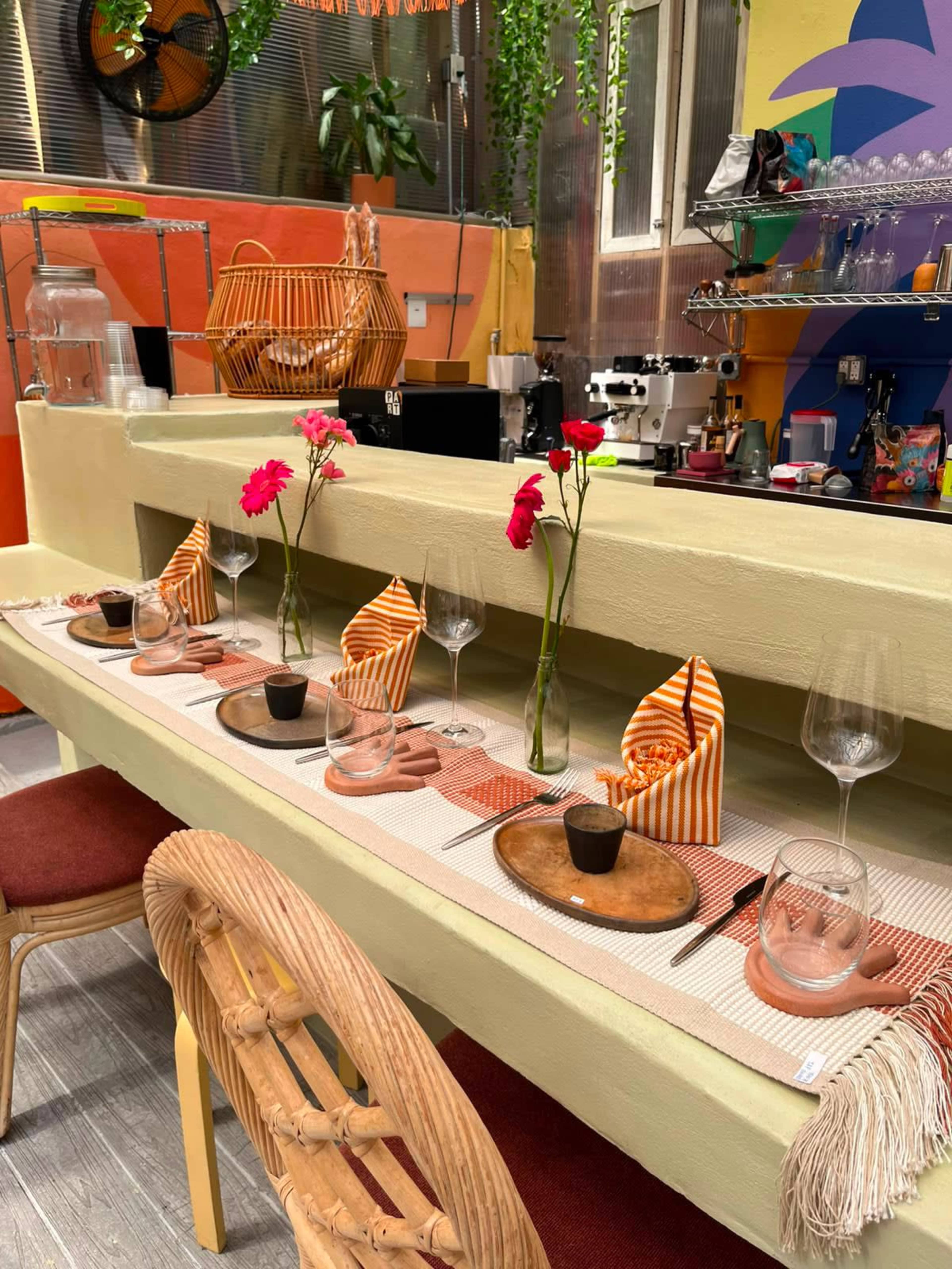 A long dining table is set with plates, glasses, and flower vases, surrounded by colorful decor and wicker chairs.