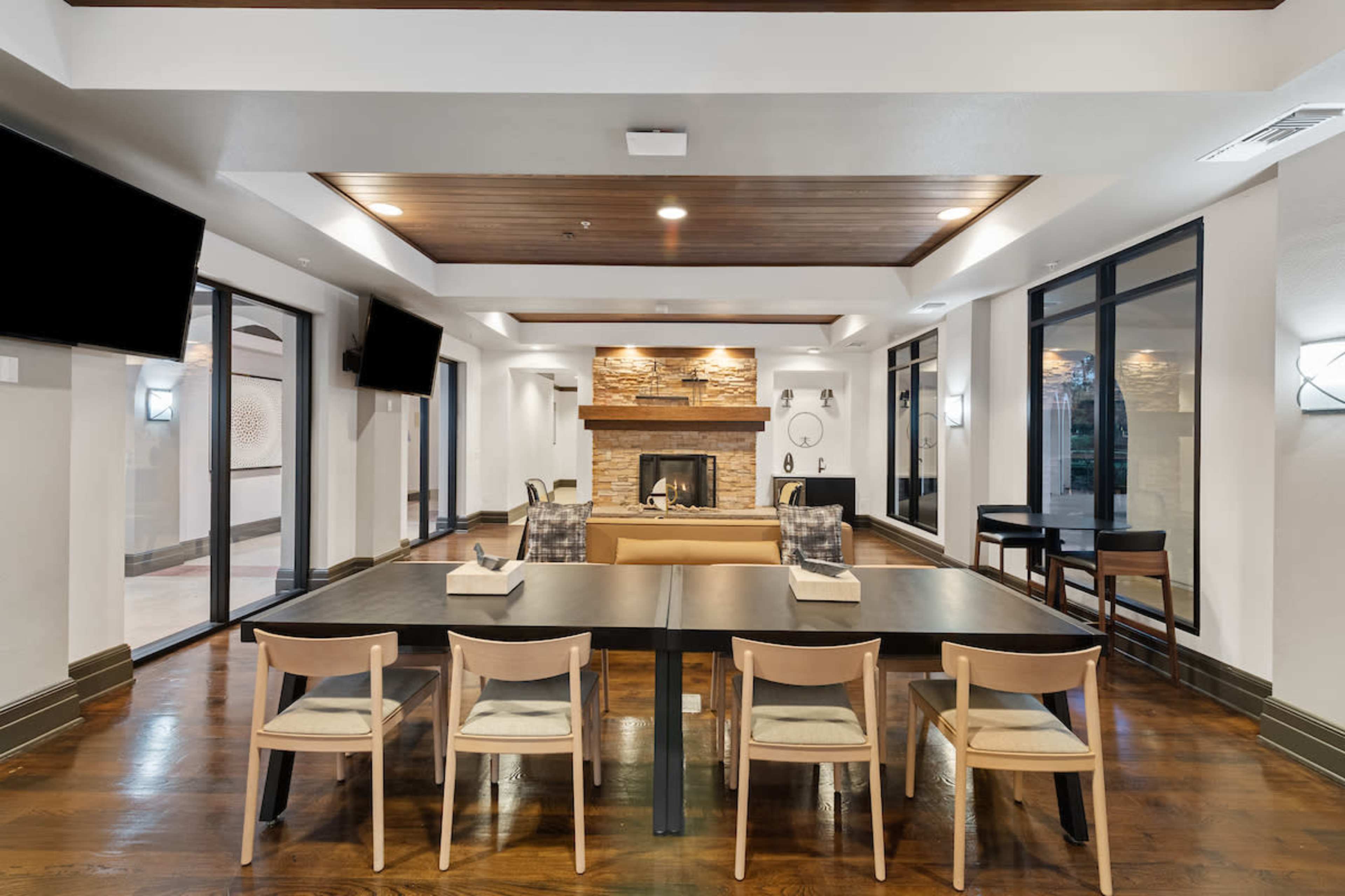 A modern lounge area featuring a wooden ceiling, a stone fireplace, seating arrangements, and large windows.