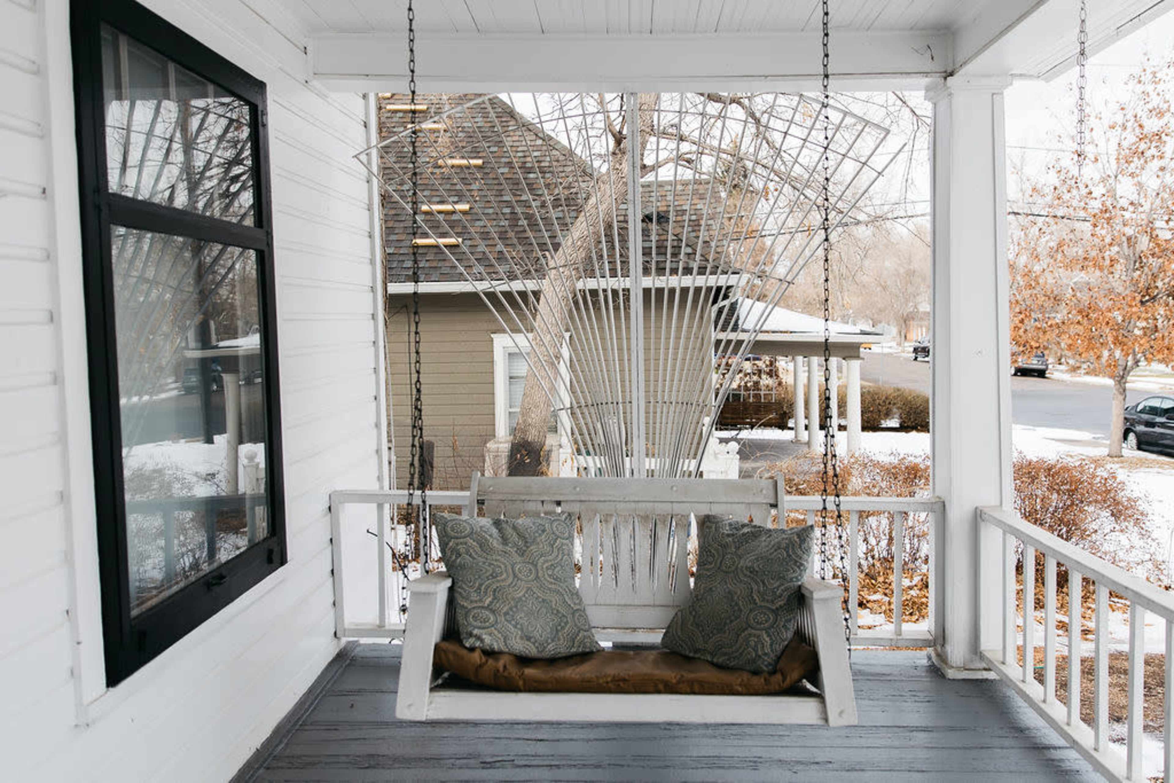 A wooden porch swing with decorative cushions hangs from a porch overlooking a snowy yard and street.