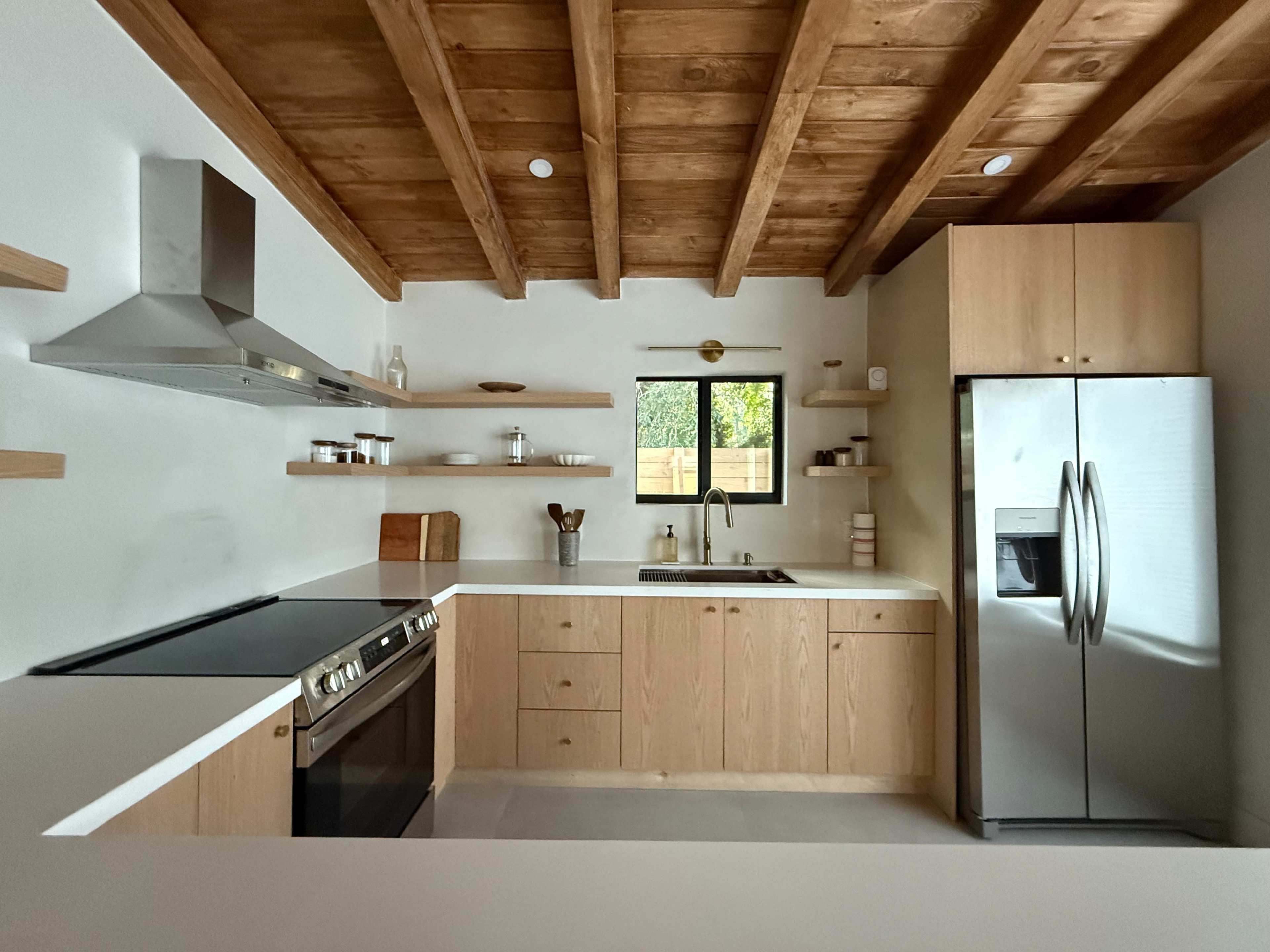 A modern kitchen features wood-beamed ceilings, light wood cabinetry, stainless steel appliances, and minimal open shelving.