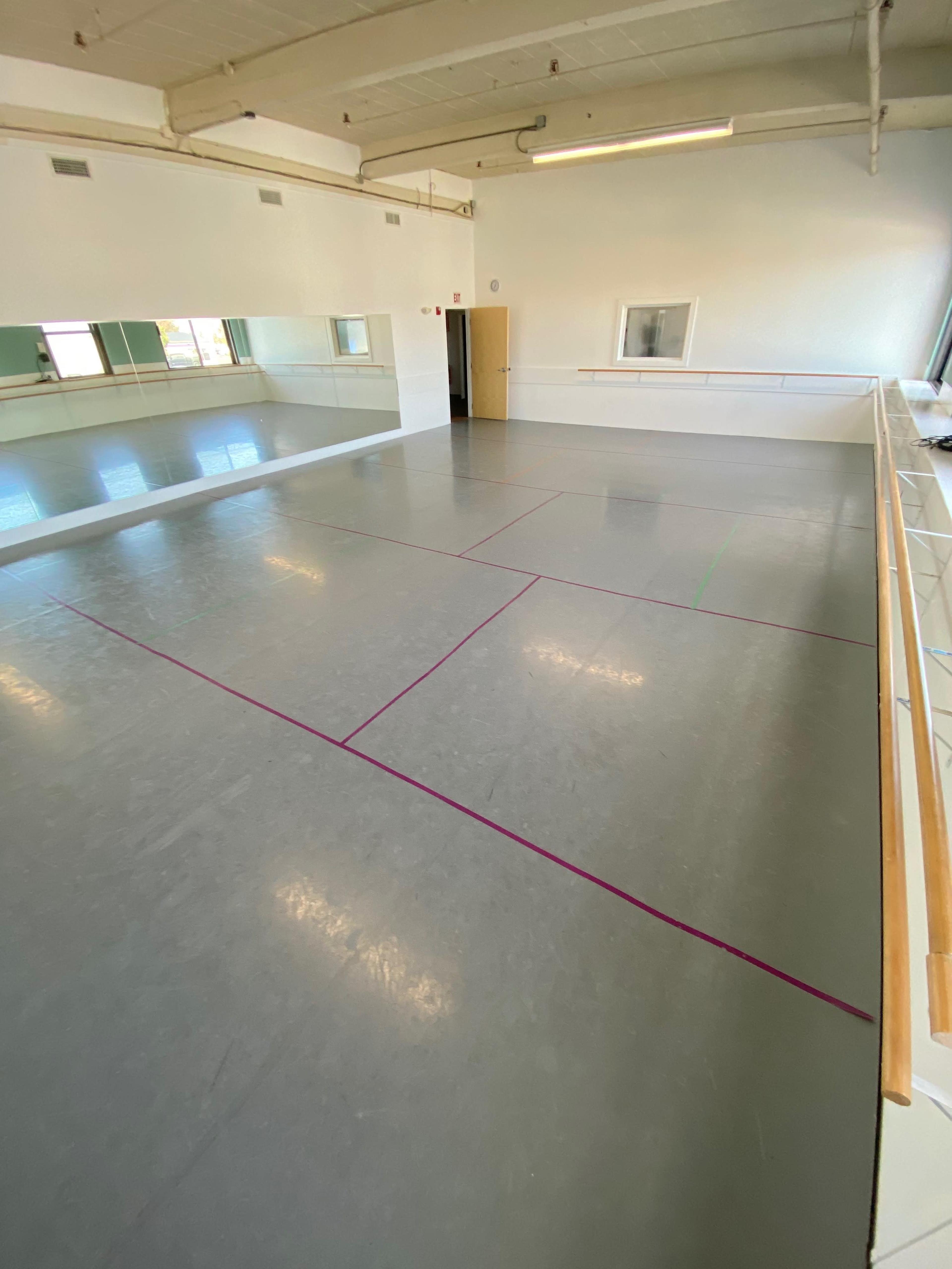 A spacious dance studio with a gray floor marked by colored lines and large mirrors on one wall.