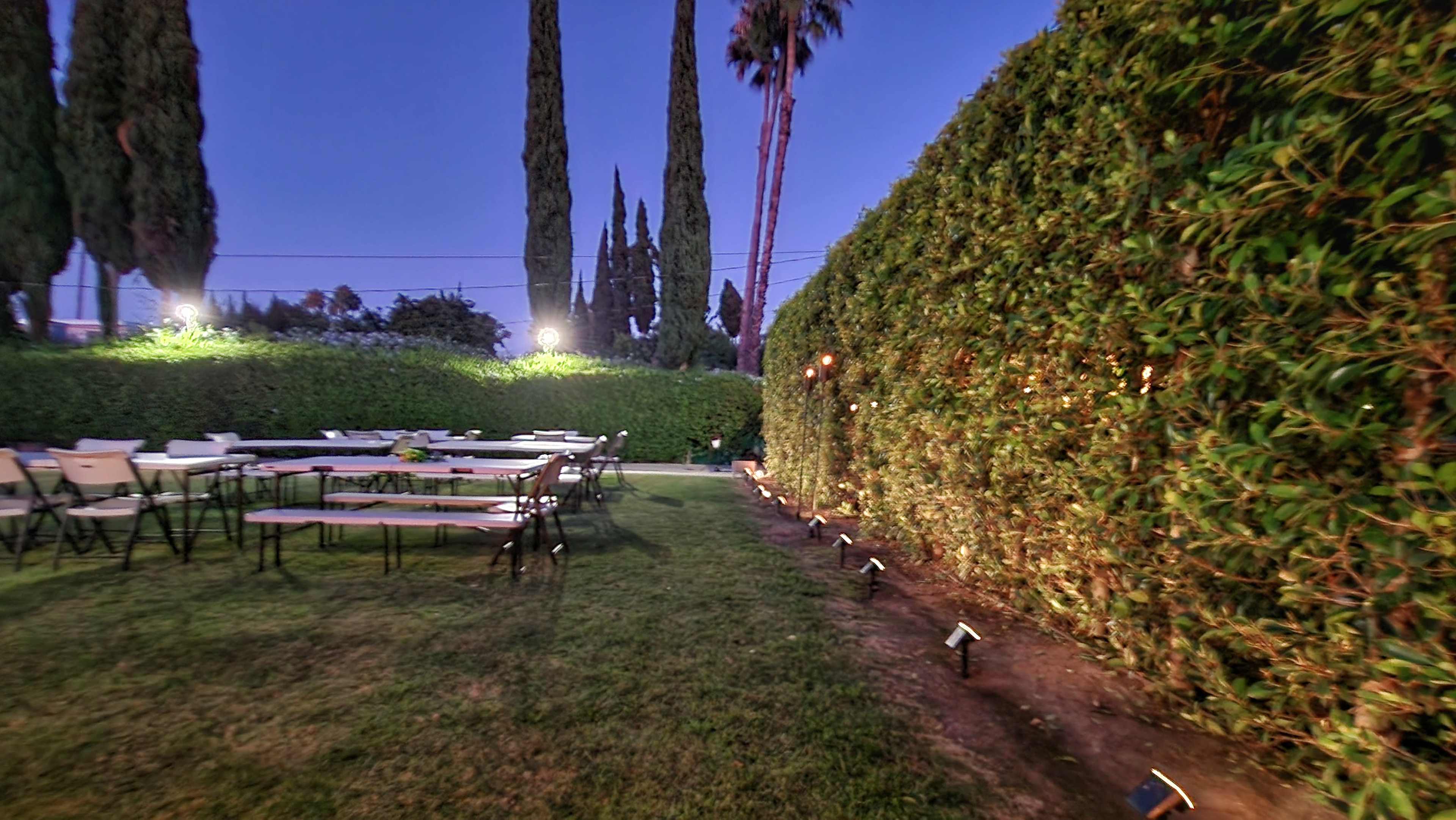 The scene shows an outdoor area with several long tables set up on the grass, surrounded by tall hedges and palm trees, illuminated by string lights at dusk.