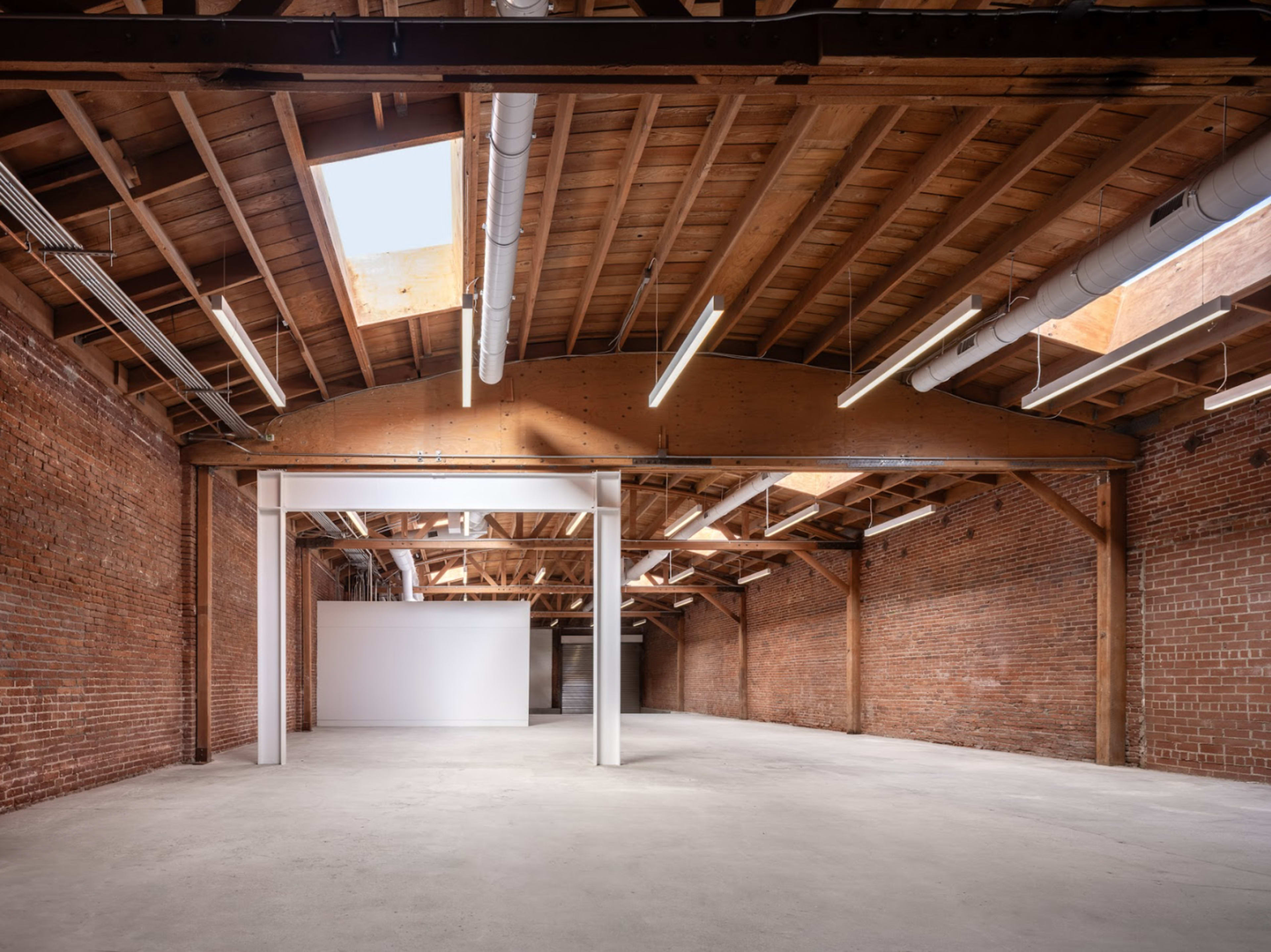 A spacious, empty warehouse with brick walls, wooden beams, and skylights illuminating the interior.