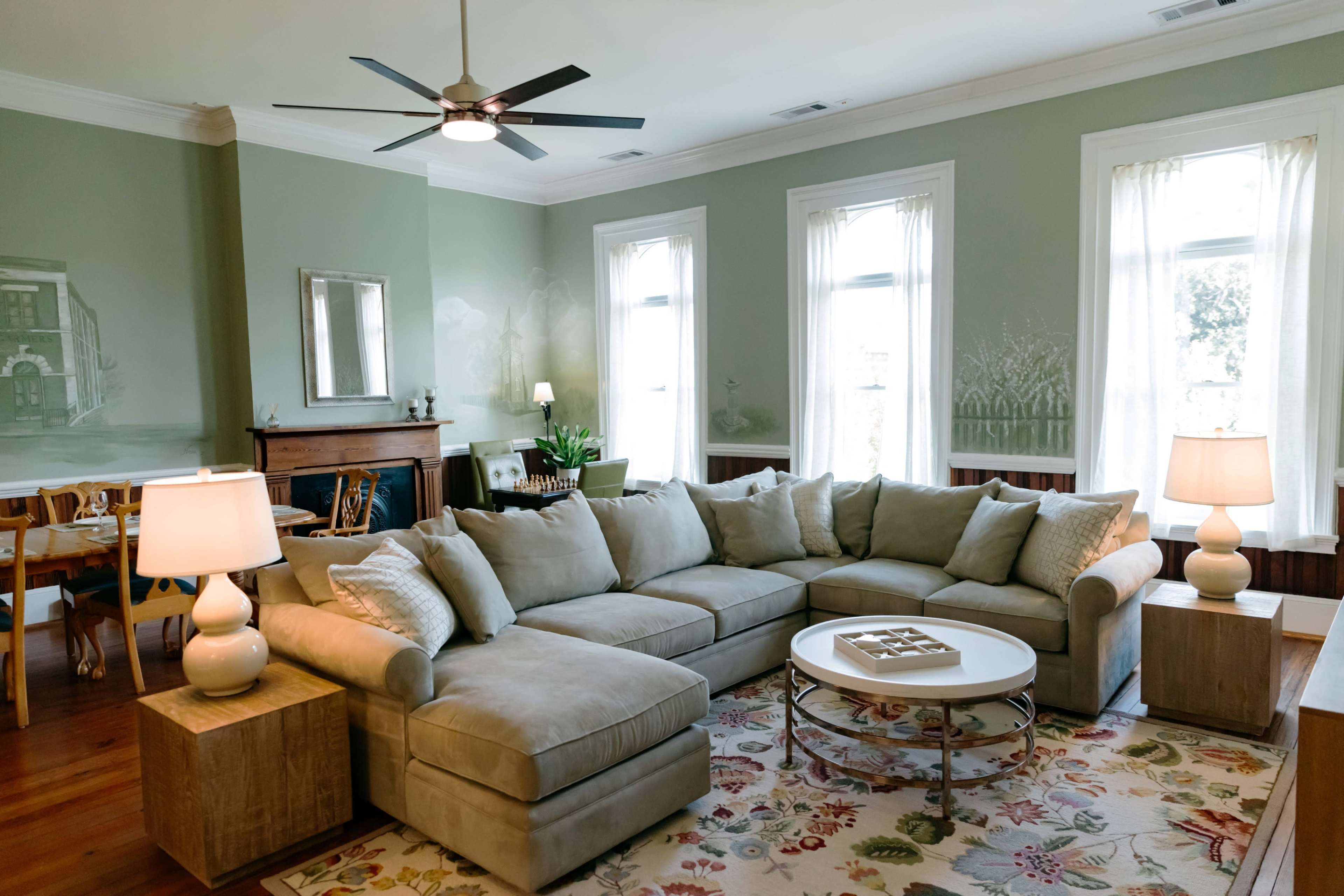 The living room features a large sectional sofa, a round coffee table, and white curtains framing the windows.