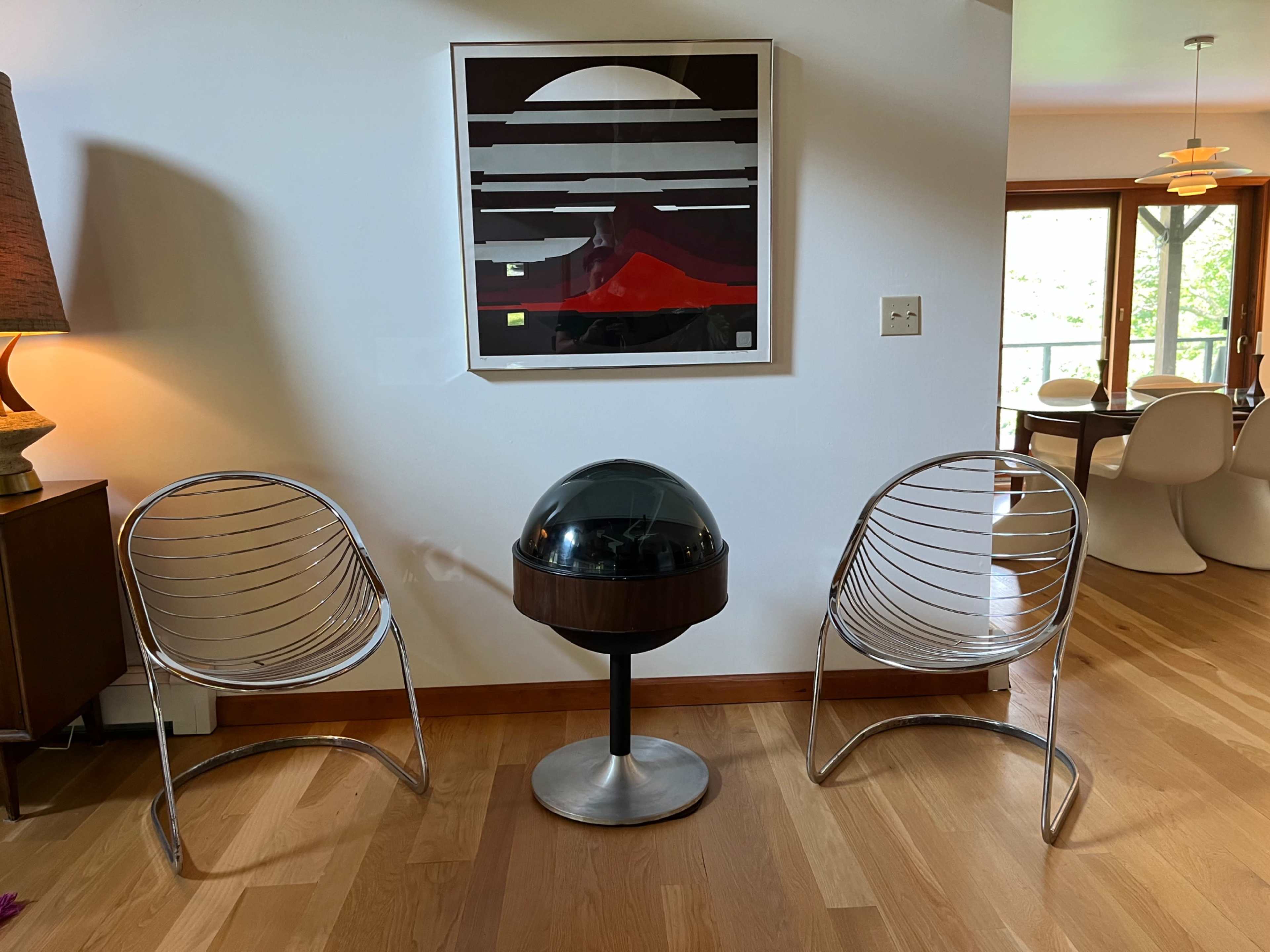 The image shows two modern wire chairs facing a round table with a black dome on top, against a backdrop of a wall featuring an abstract artwork.