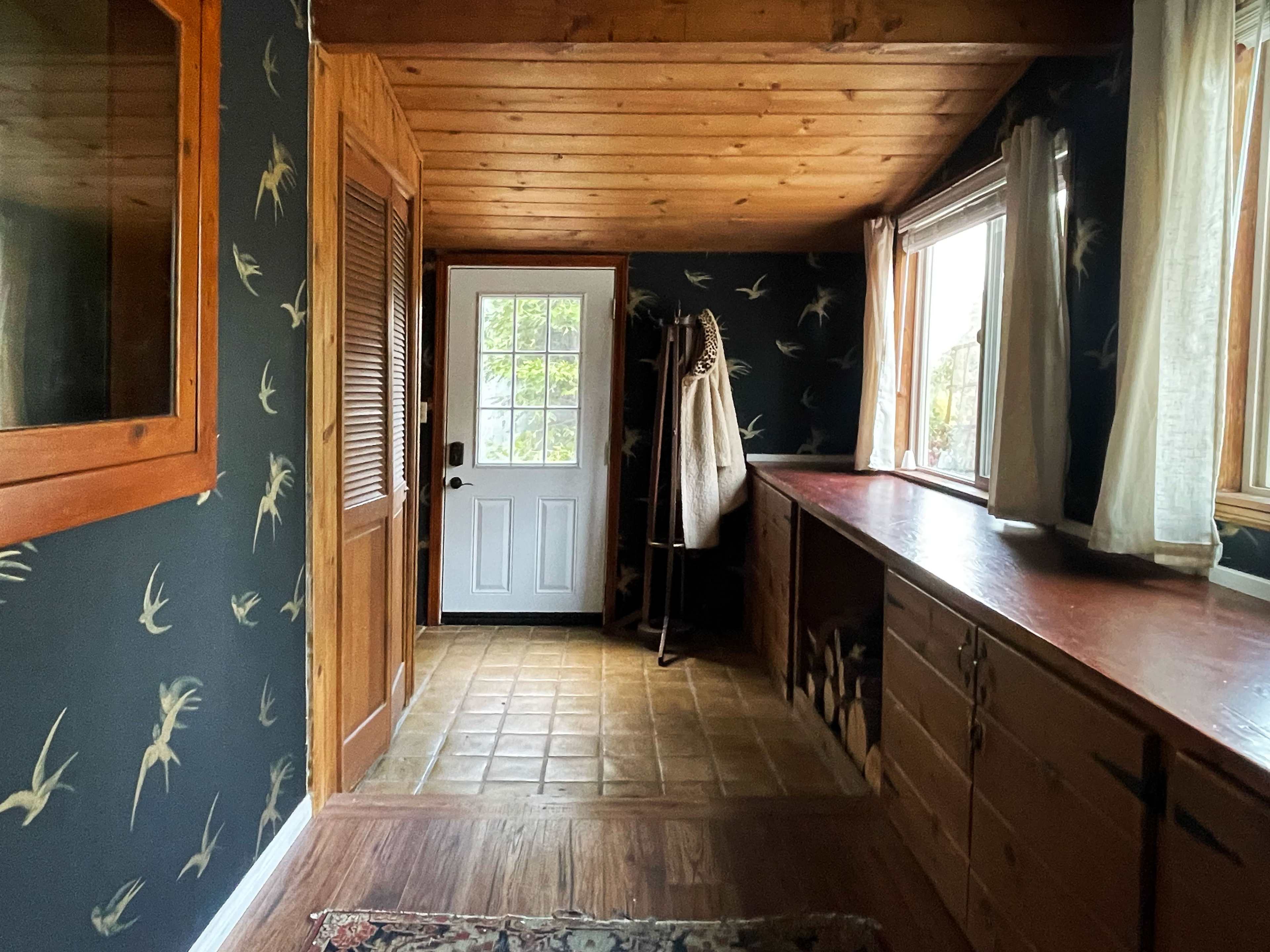 A hallway with wooden walls and a door leading outside, featuring a patterned wallpaper, a window, and a countertop along one side.