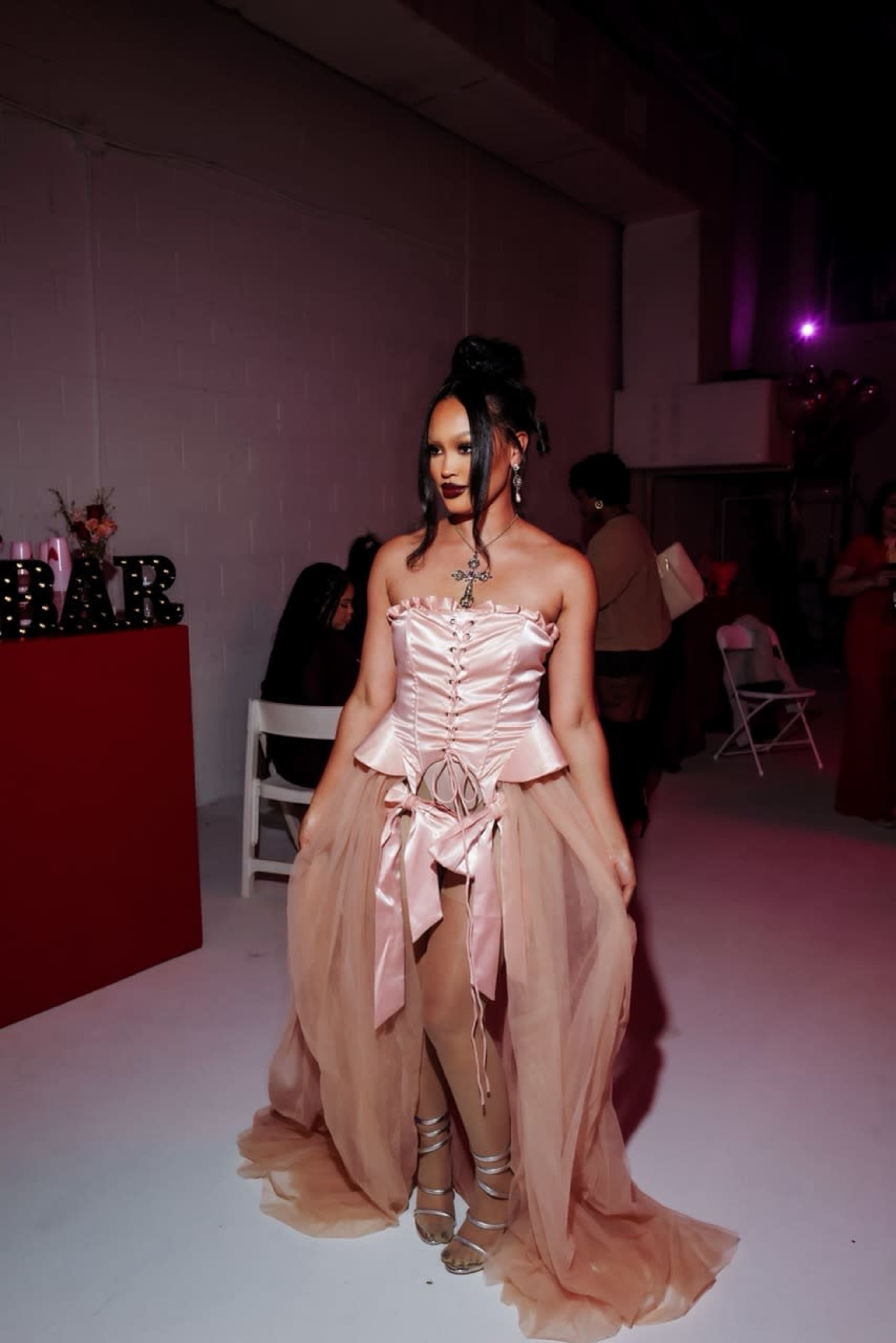 A woman in a pink corset dress with a sheer overlay stands in a brightly lit event space.