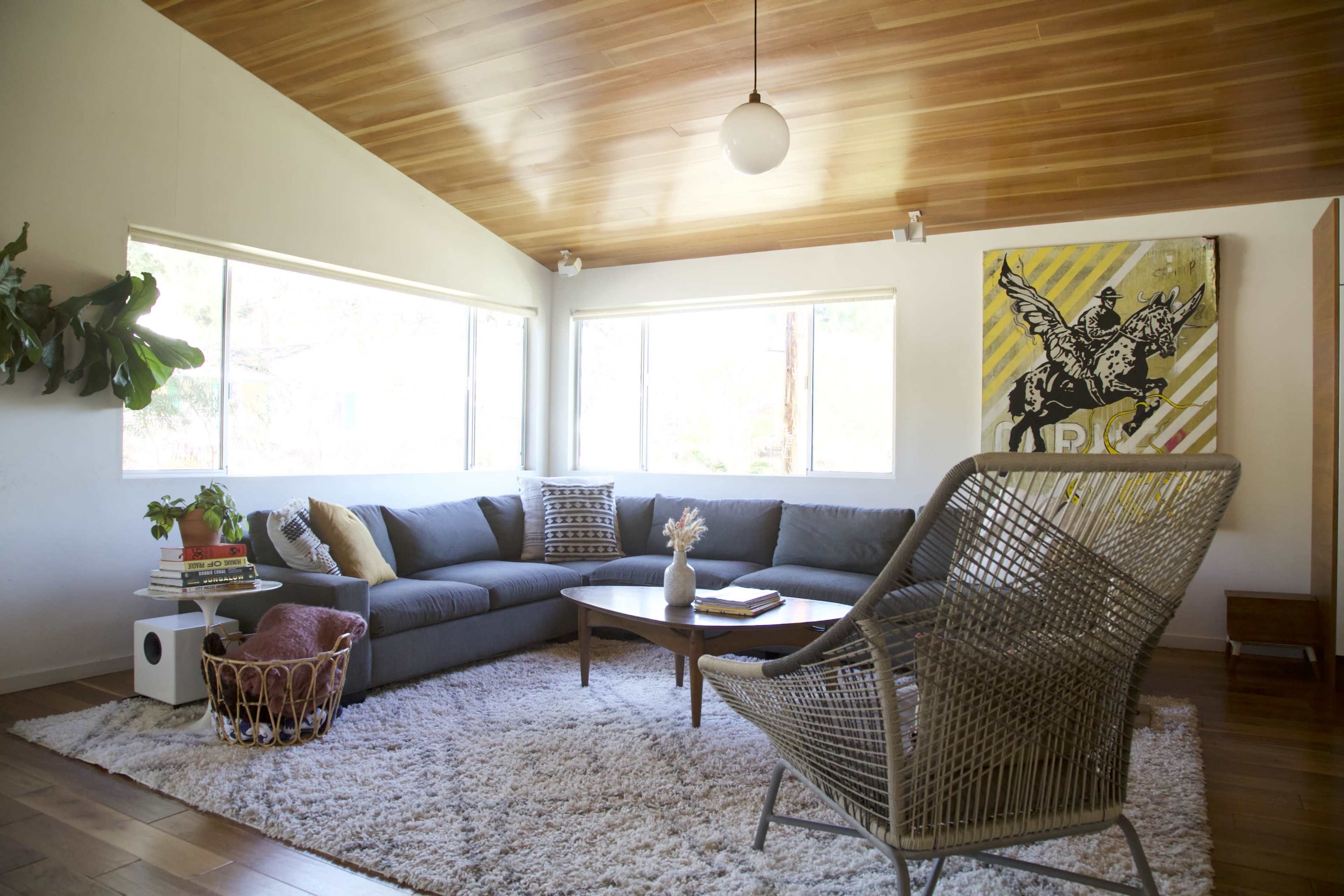 A modern living room features a large gray sectional sofa, a round wooden coffee table, and a wicker chair, with natural light streaming in through two windows.