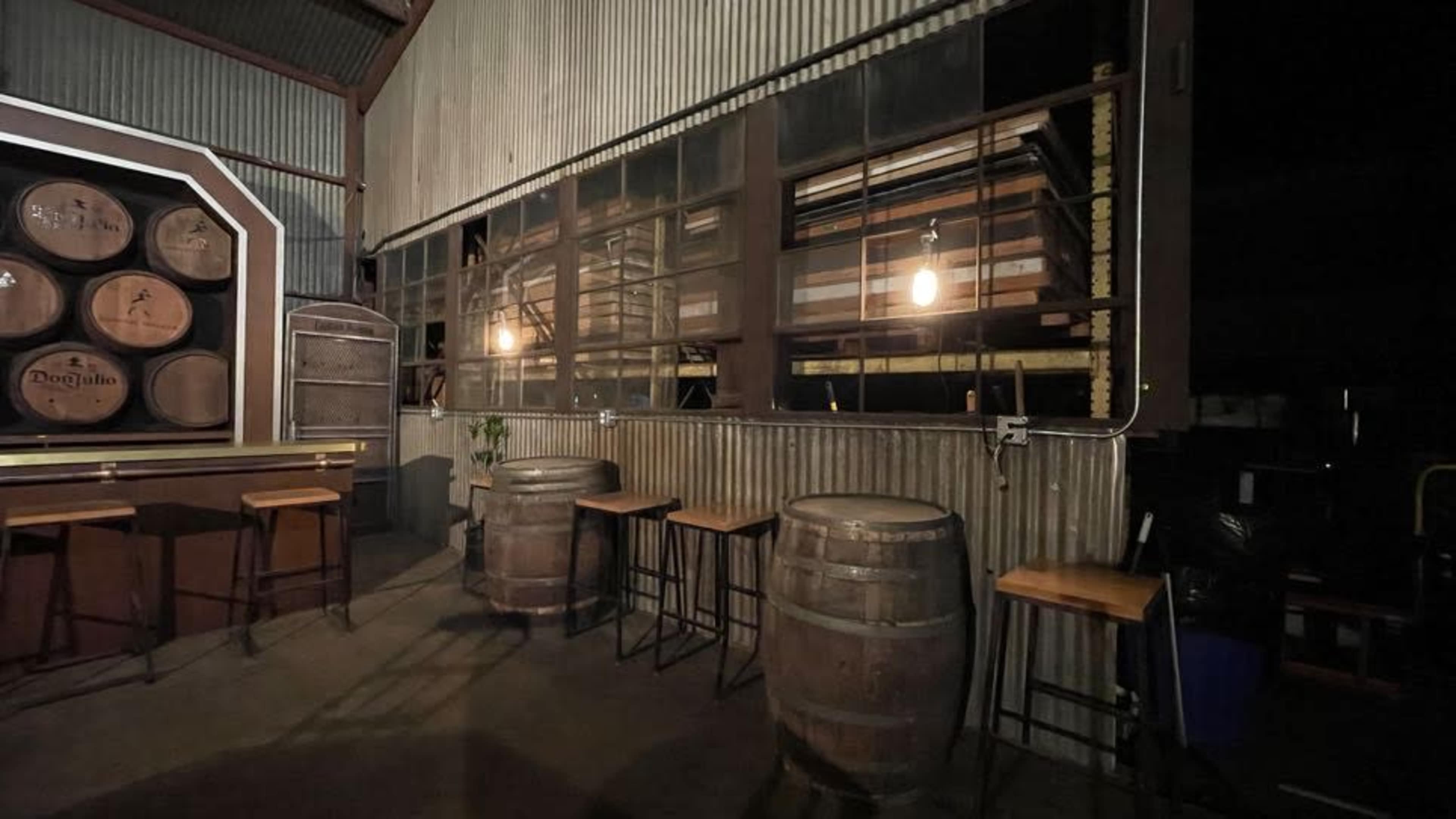 The scene shows a dimly lit interior of a bar with wooden barrels and stools against a wall lined with large windows.