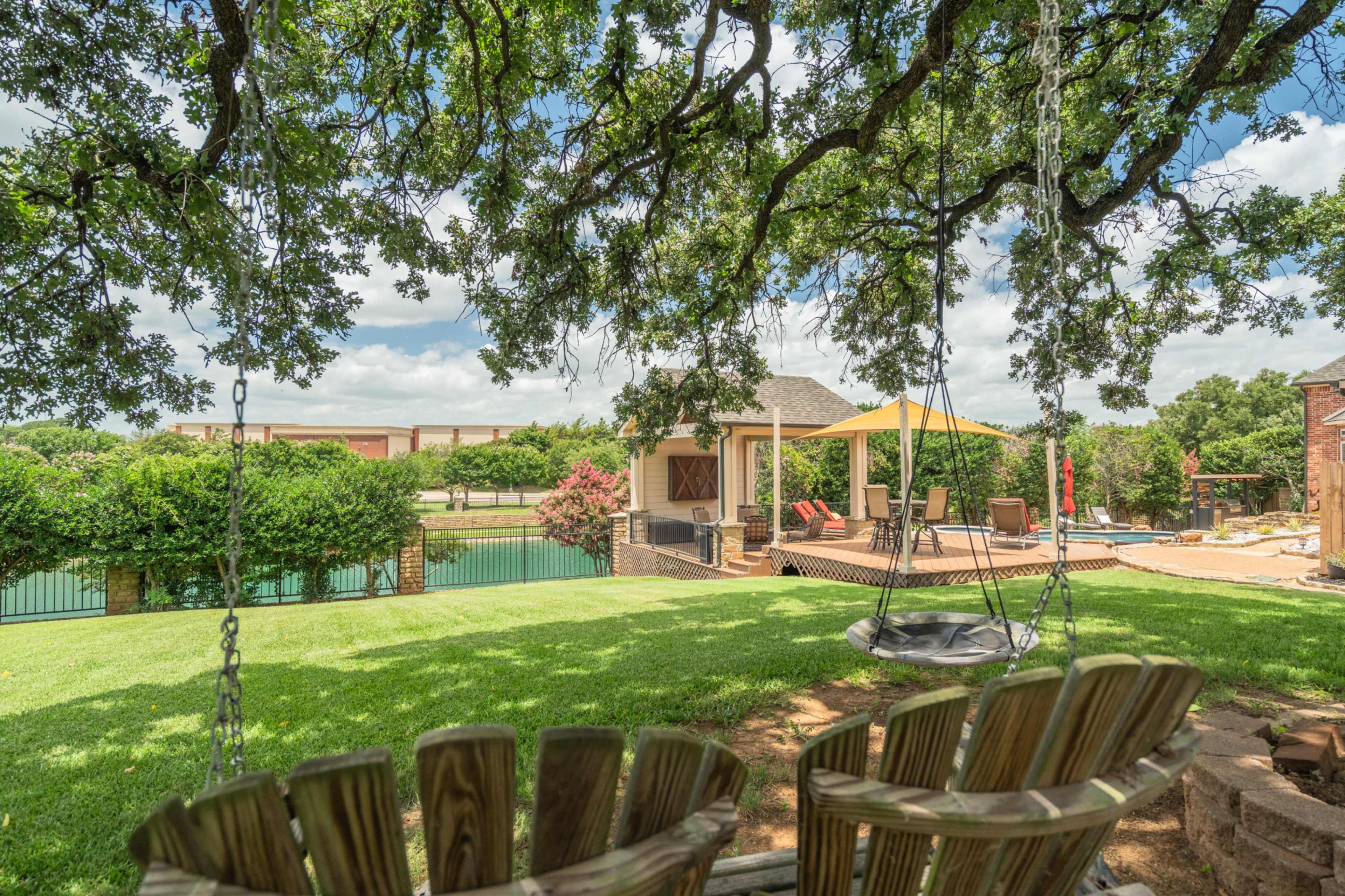 A backyard scene features a swing under a large tree, overlooking a pool, patio area, and a gazebo.