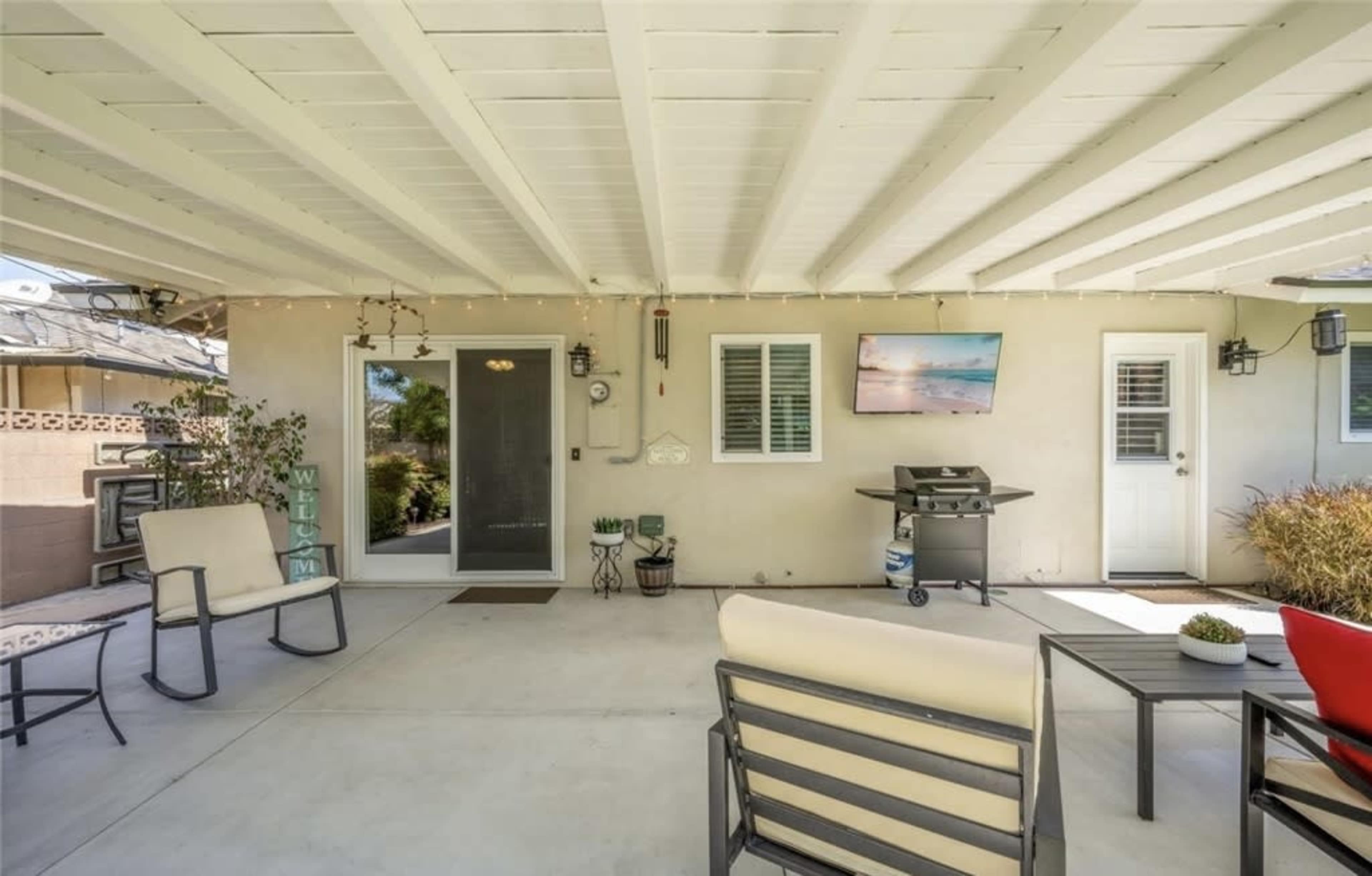 The image shows a covered patio area with outdoor seating, a grill, and decorative lighting.