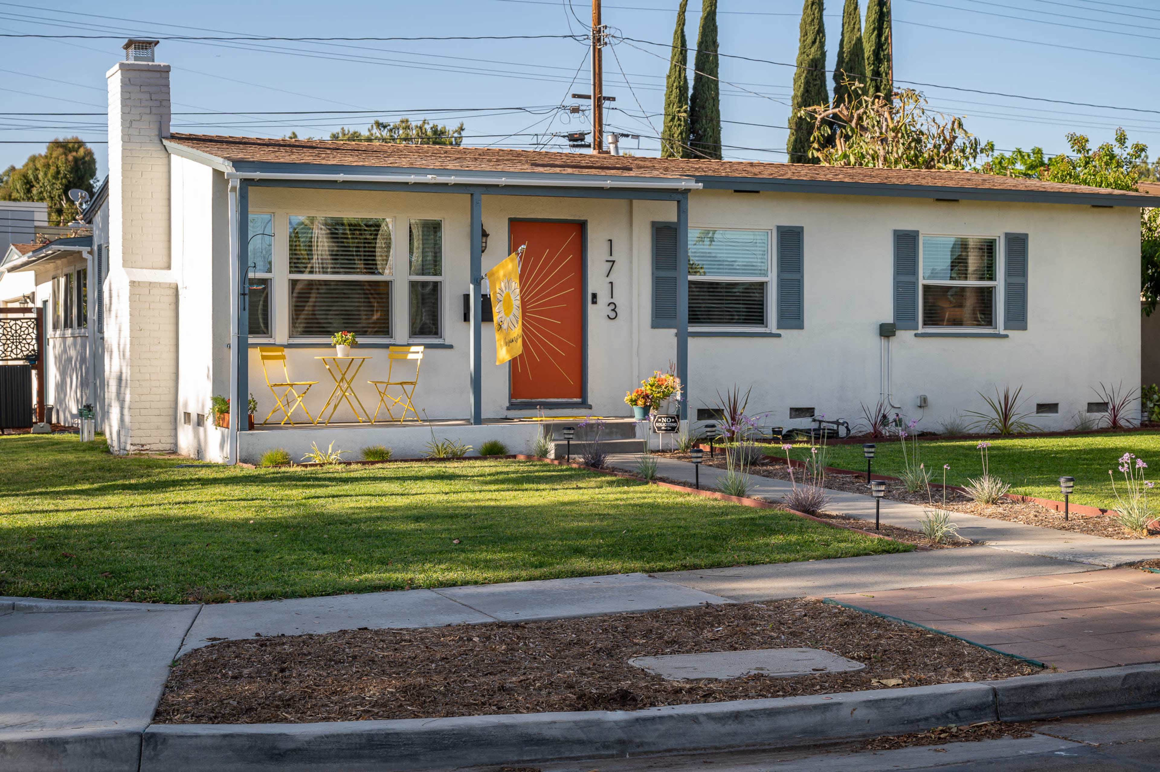A single-story, light-colored house with a vibrant orange door, yellow furniture on the porch, and landscaped grass in front.