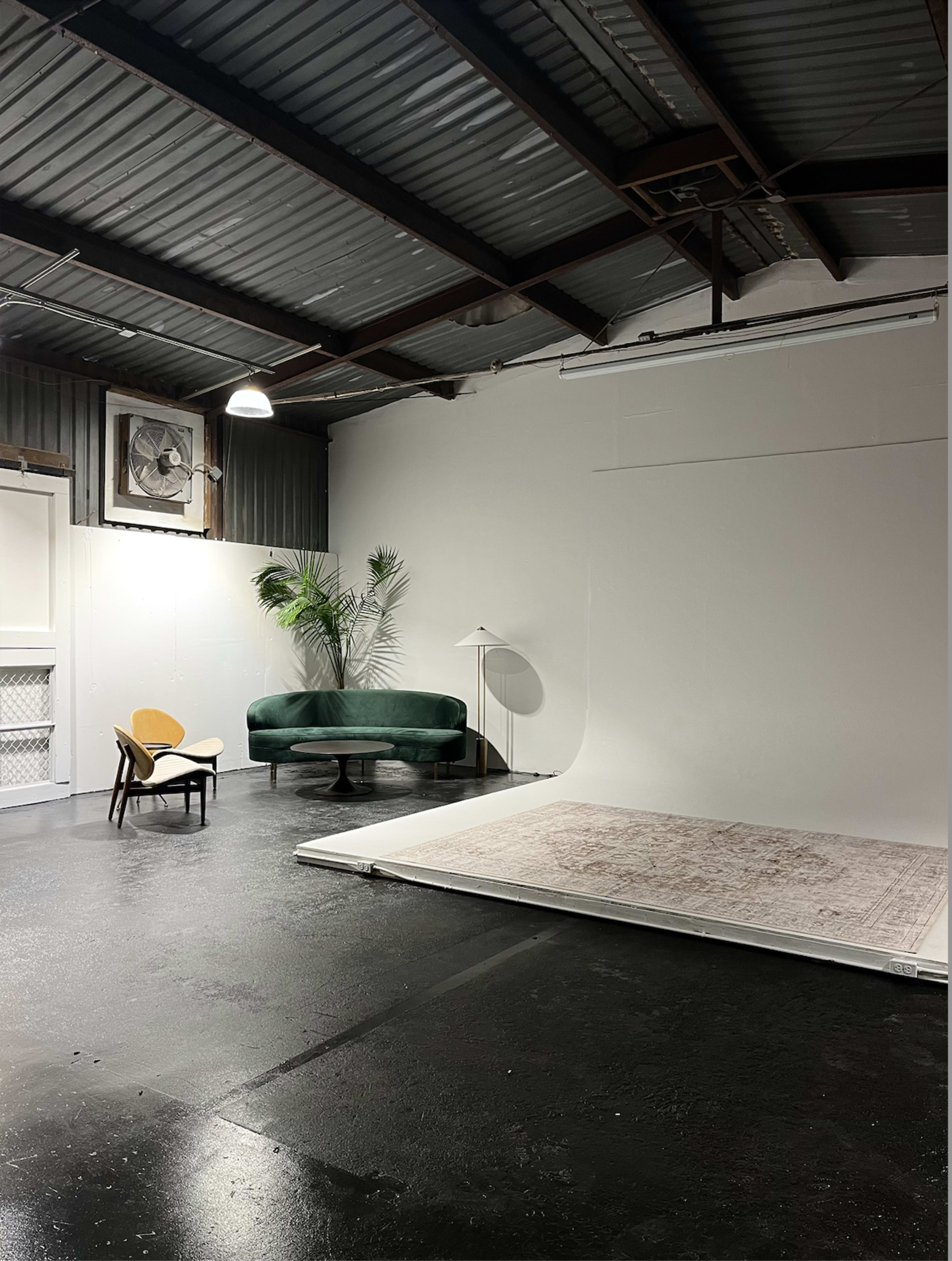 A spacious studio with a large white backdrop, a green sofa, a black armchair, and a rug on a polished black floor.