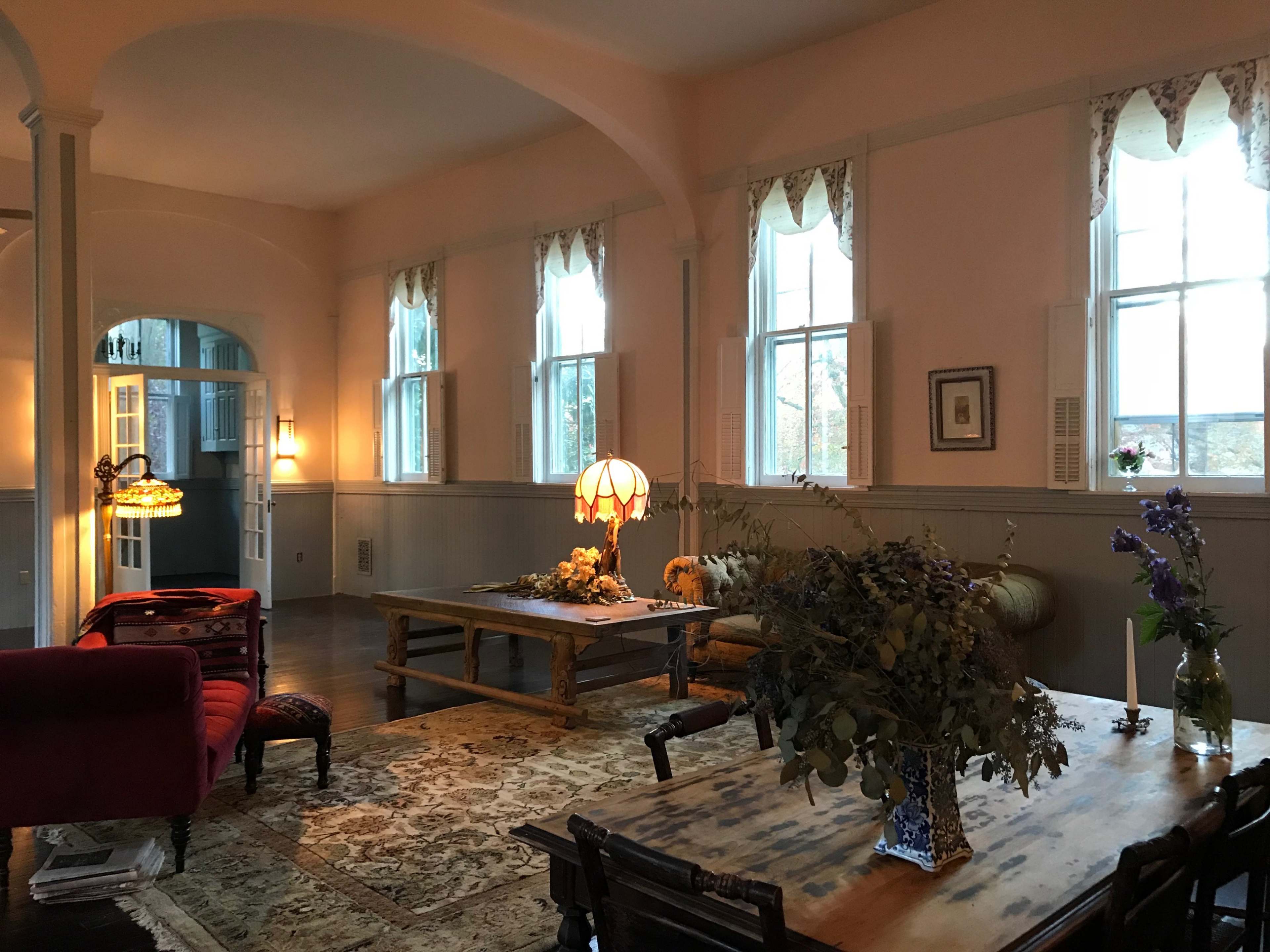 A spacious, well-lit living area with a red sofa, wooden tables, and large windows adorned with curtains.