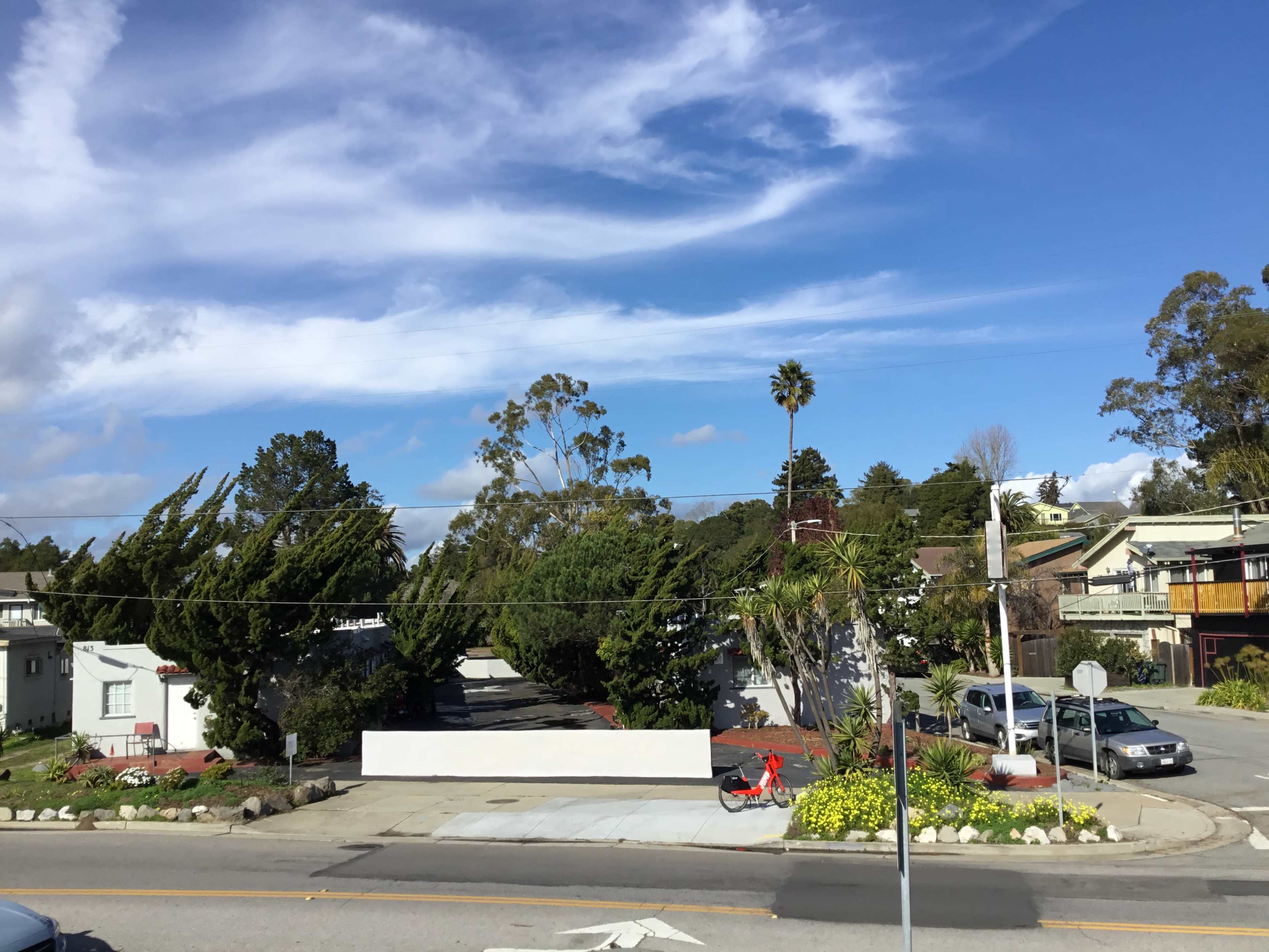 The image shows a residential area with trees leaning in the wind, a blue sky with clouds, and a partially visible street corner.