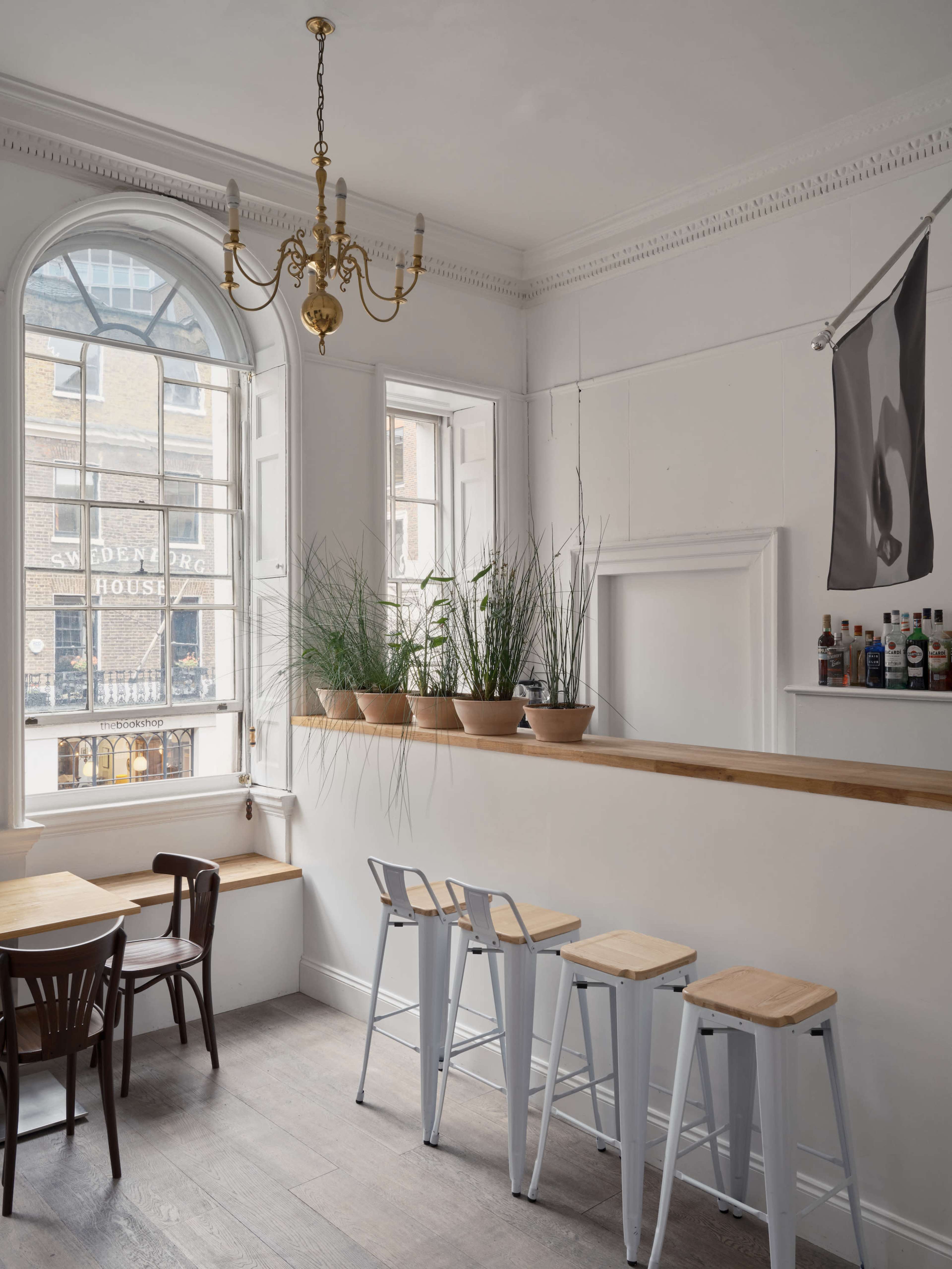 A minimalistic cafe interior with wooden bar stools, potted plants on the windowsill, and a large arched window revealing a street view.