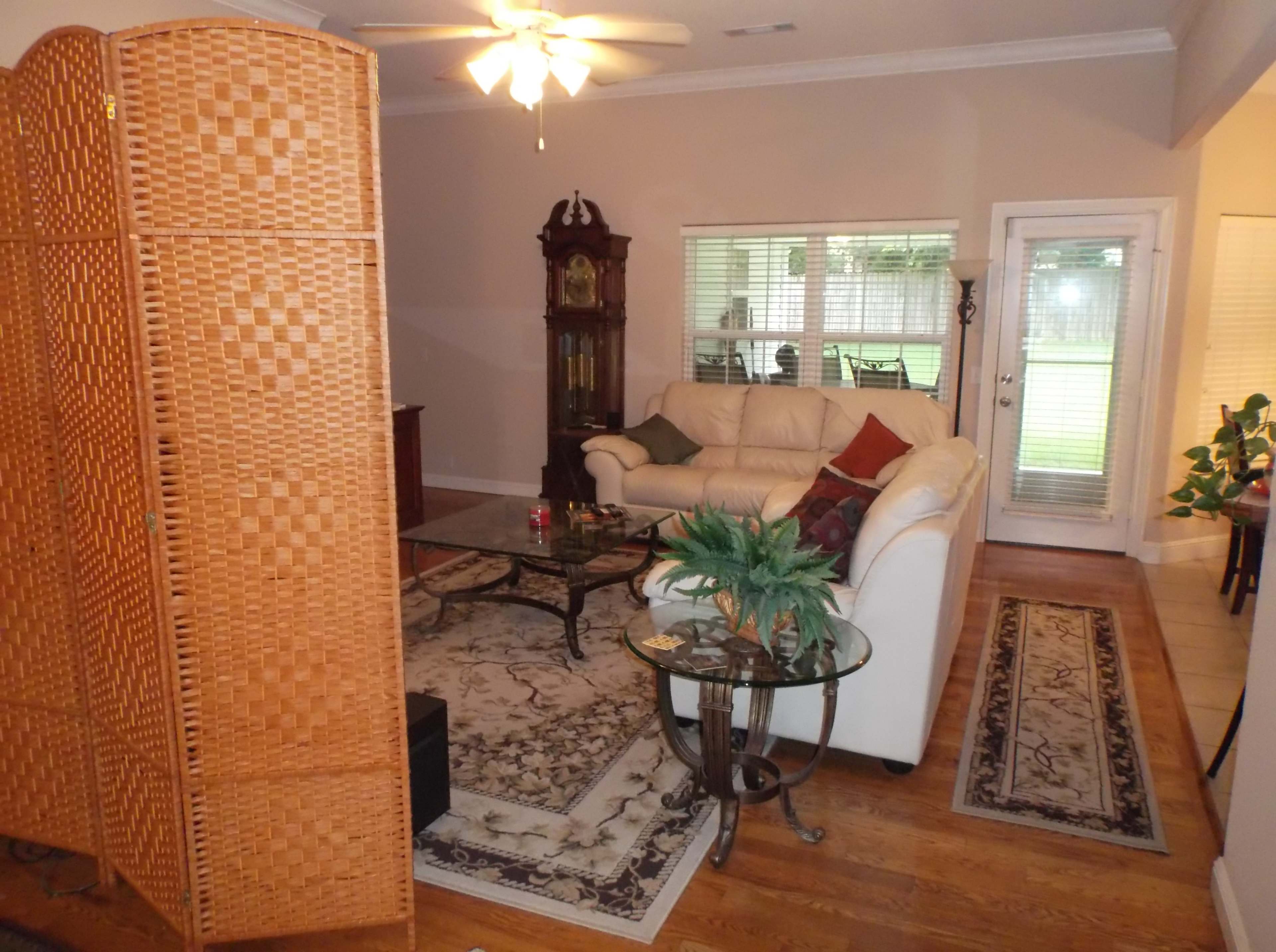 A living room featuring a beige sofa, a glass coffee table, a decorative plant, and a wooden screen separating spaces, with large windows providing natural light.