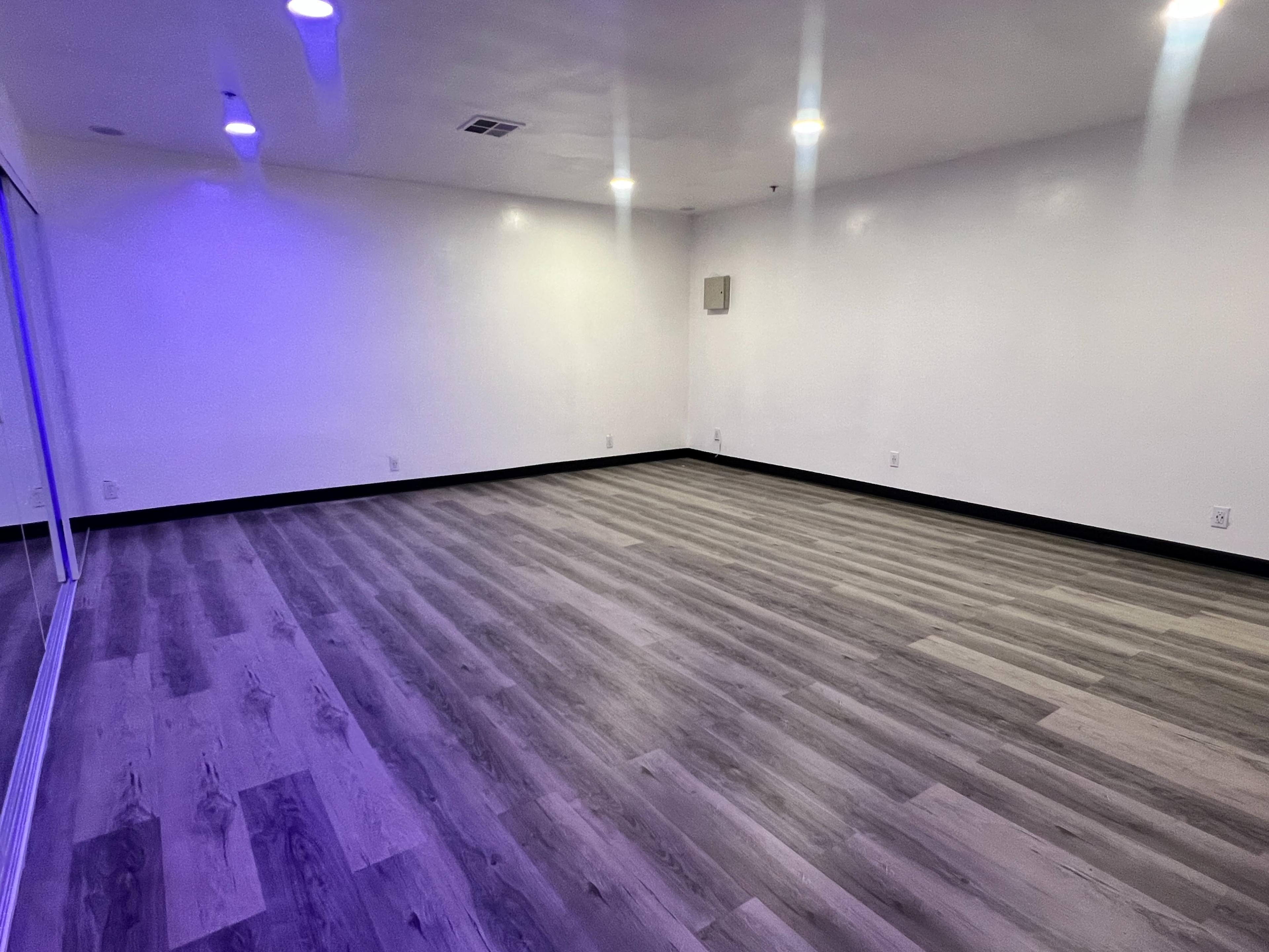 The image shows a vacant room with wooden flooring and white walls, illuminated by overhead lights.