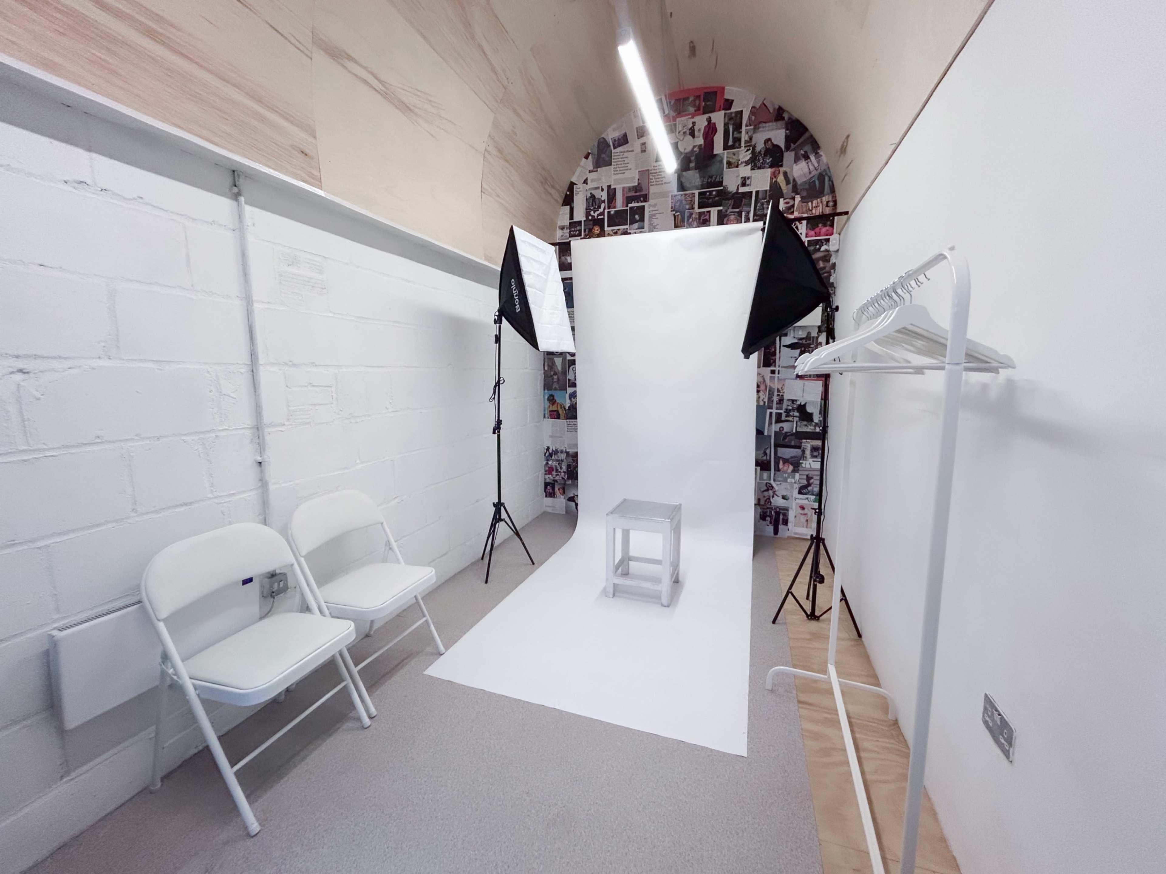 The image shows a minimalist photography studio featuring a white backdrop, two softbox lights, a stool, and two folding chairs.