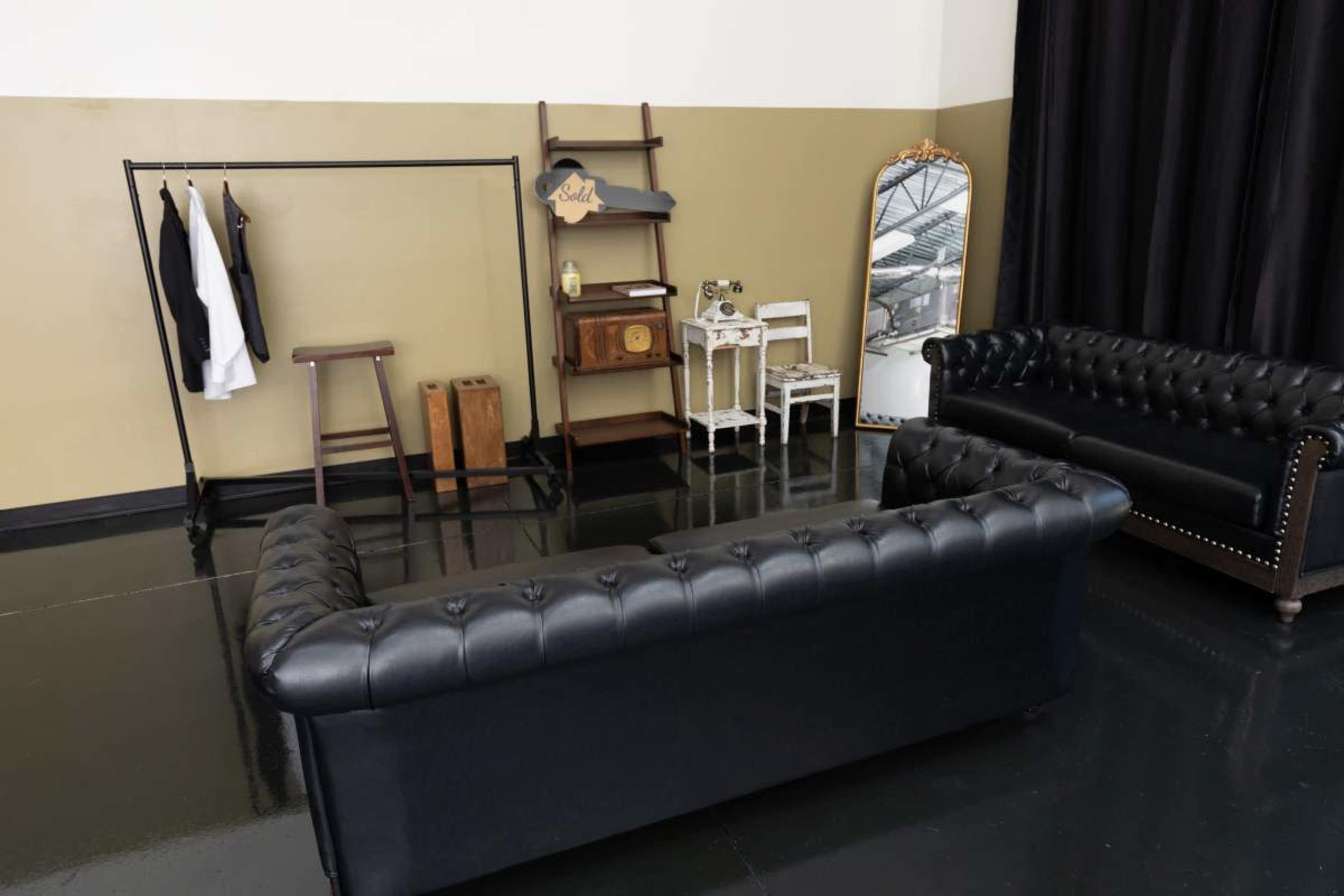 A stylish interior features a black leather sofa, a clothing rack with garments, a wooden stool, and a decorative mirror against a neutral wall.