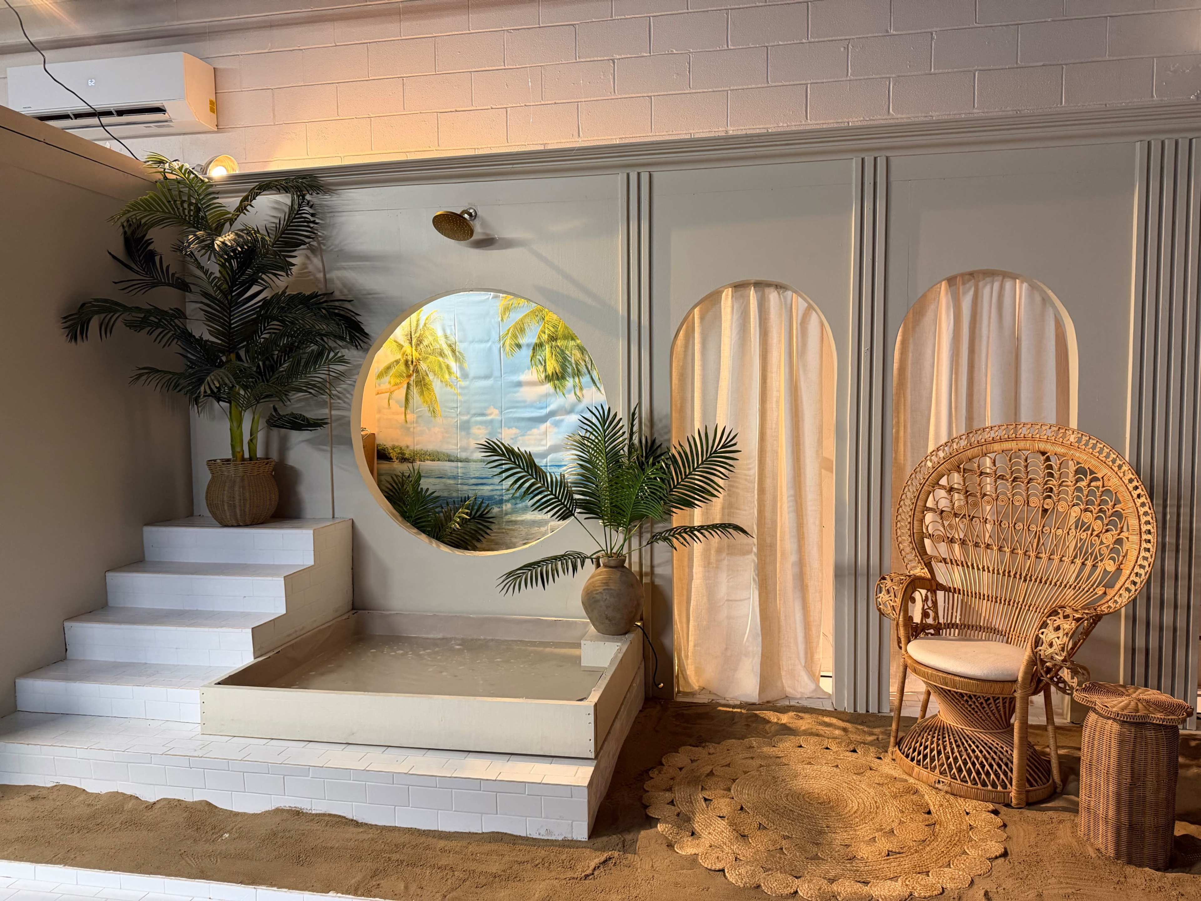 The scene features a tropical-themed interior with a rounded window depicting palm trees, surrounded by indoor plants, a rattan chair, and sand on the floor.