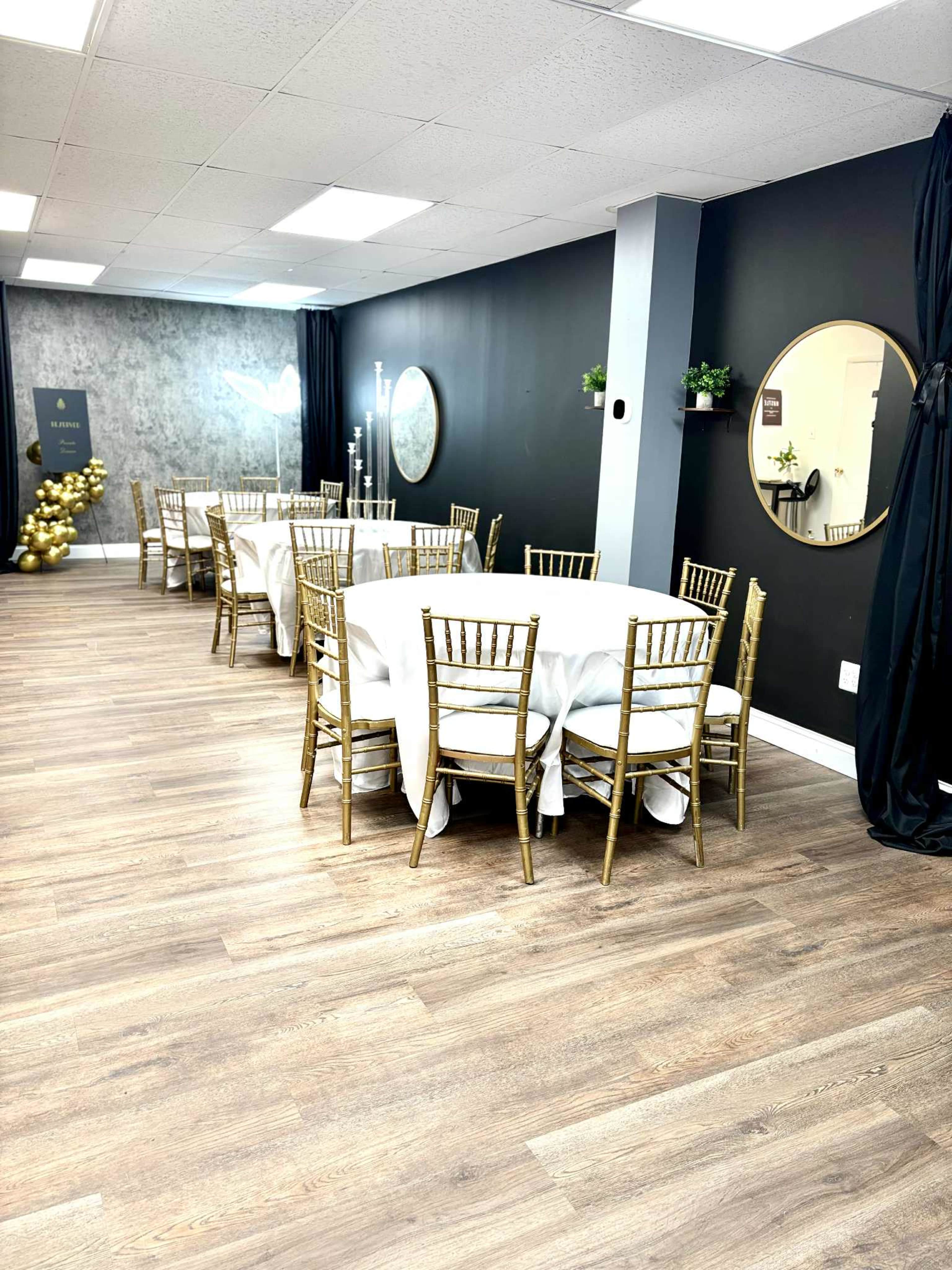 The image shows a modern event space with multiple round tables covered in white tablecloths and gold chiavari chairs, set against a dark wall and a decorative mirror.