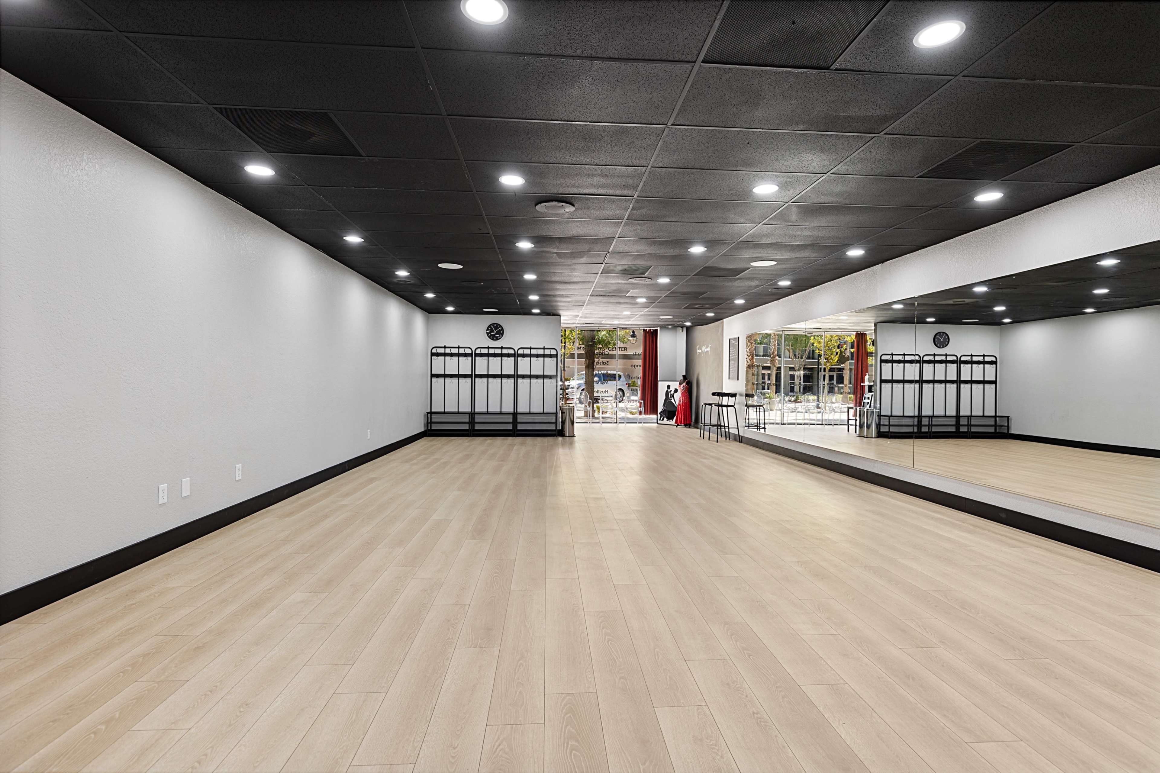 An empty dance studio with wooden flooring, mirrored walls, and a few coat racks along one side.