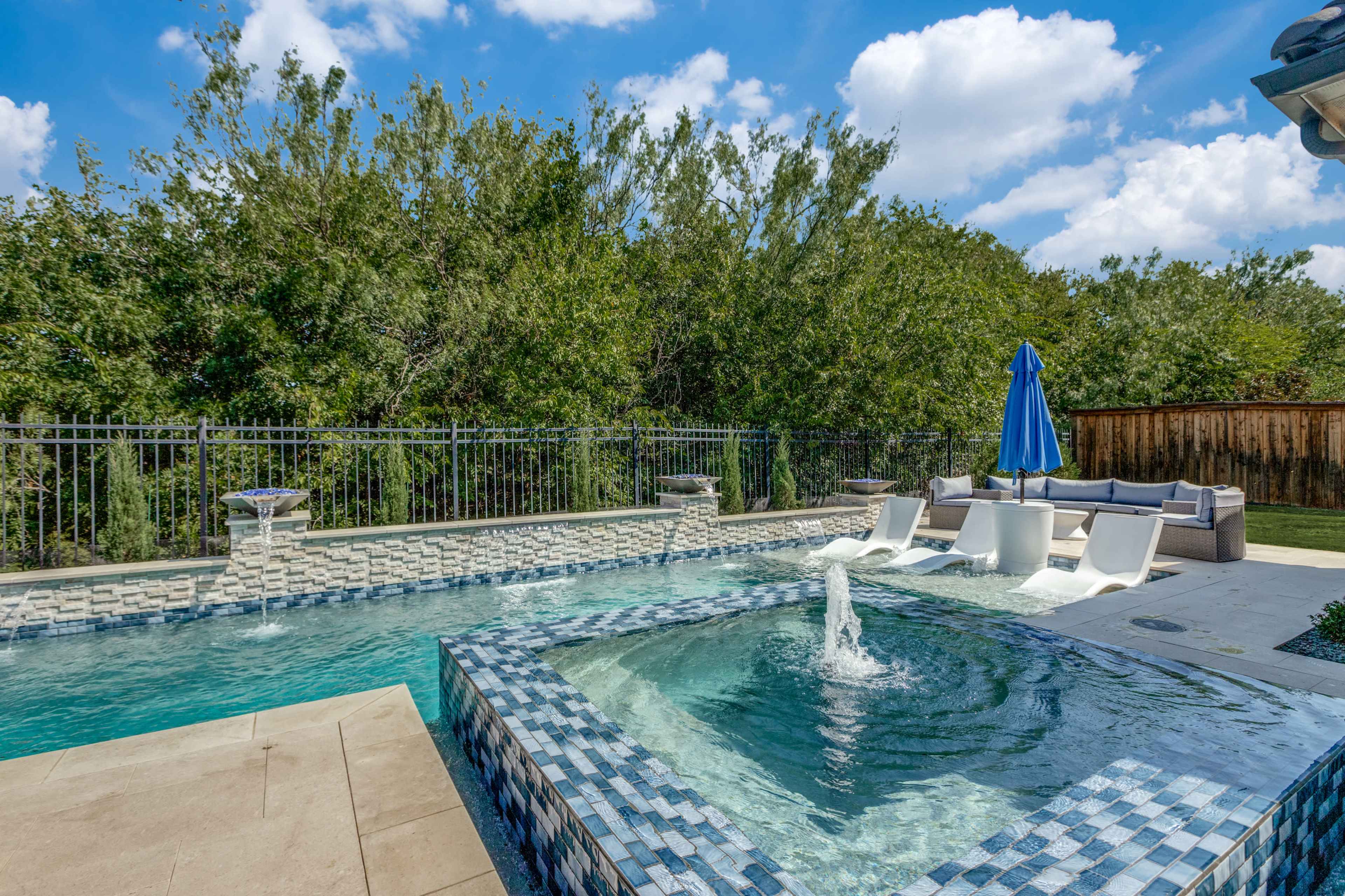 The image shows a backyard with a swimming pool, a spa area, lounge chairs, and a blue umbrella, surrounded by trees and a wooden fence.