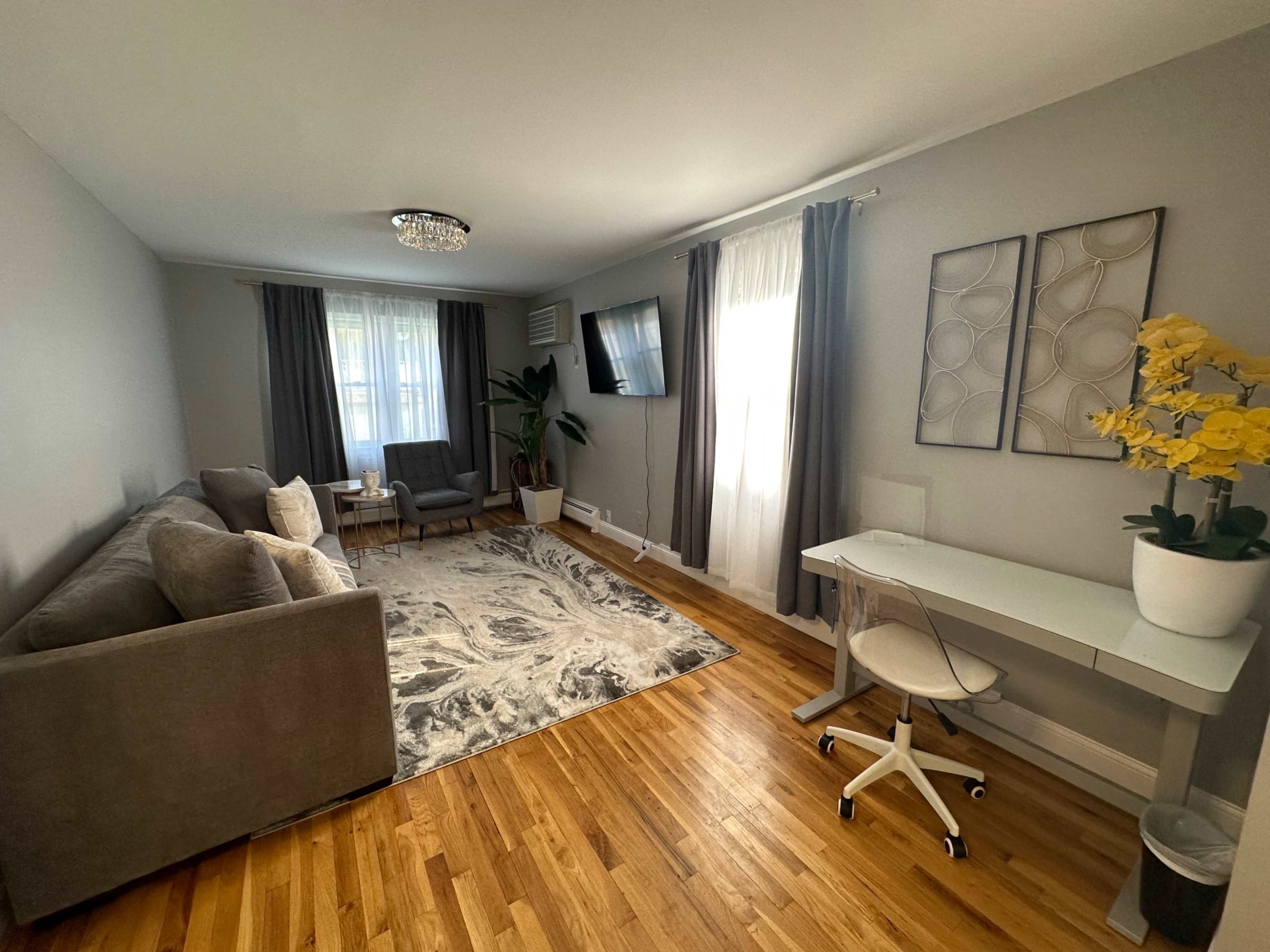 A living room with a gray sofa, a small sitting area, a desk, and a large window with light curtains.