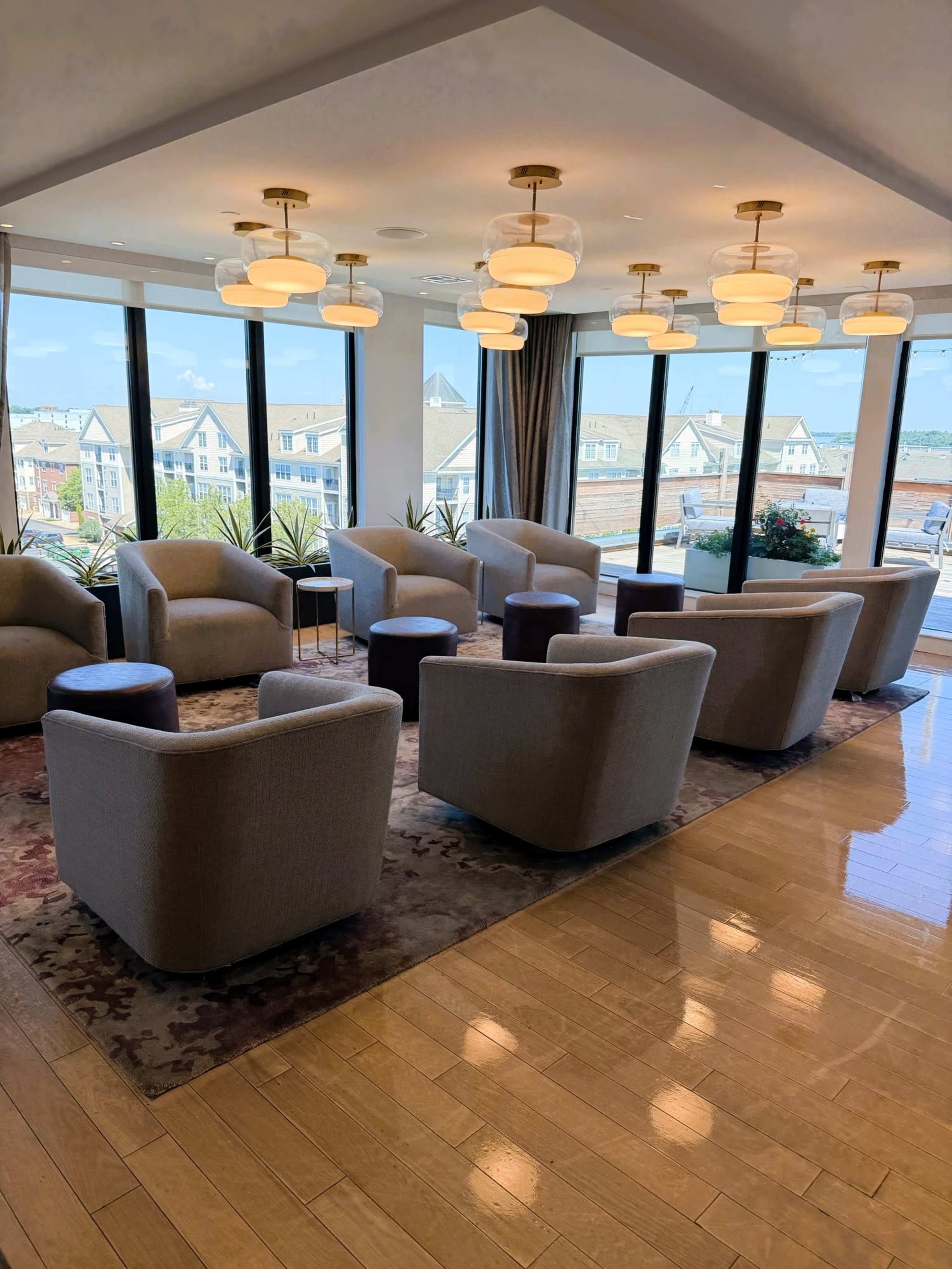 The image shows a modern lounge area with several gray armchairs arranged around small dark tables and large windows offering a view of buildings and greenery outside.