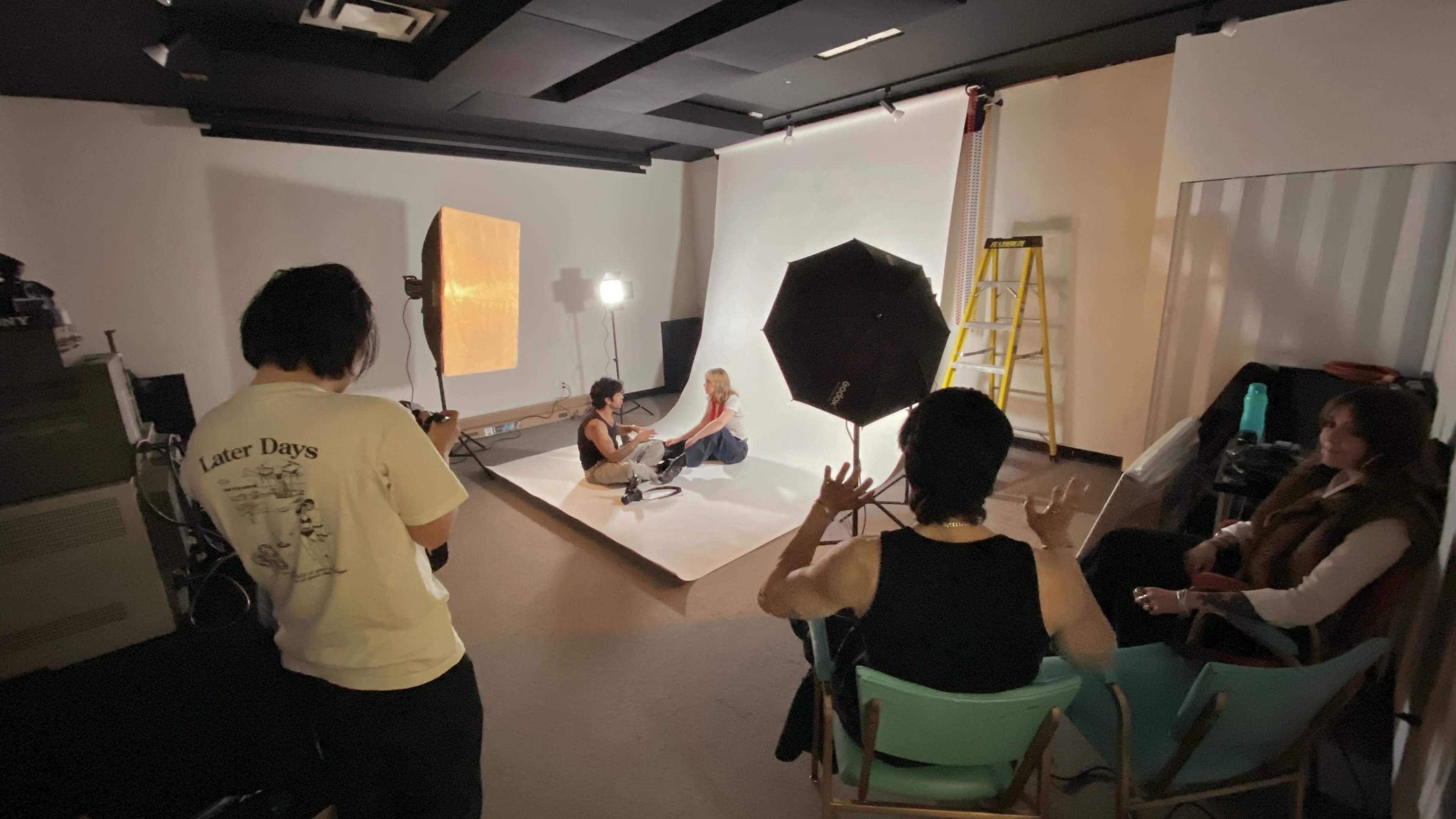 A photography studio features a model seated on a set with lights, a backdrop, and a photographer directing the scene.