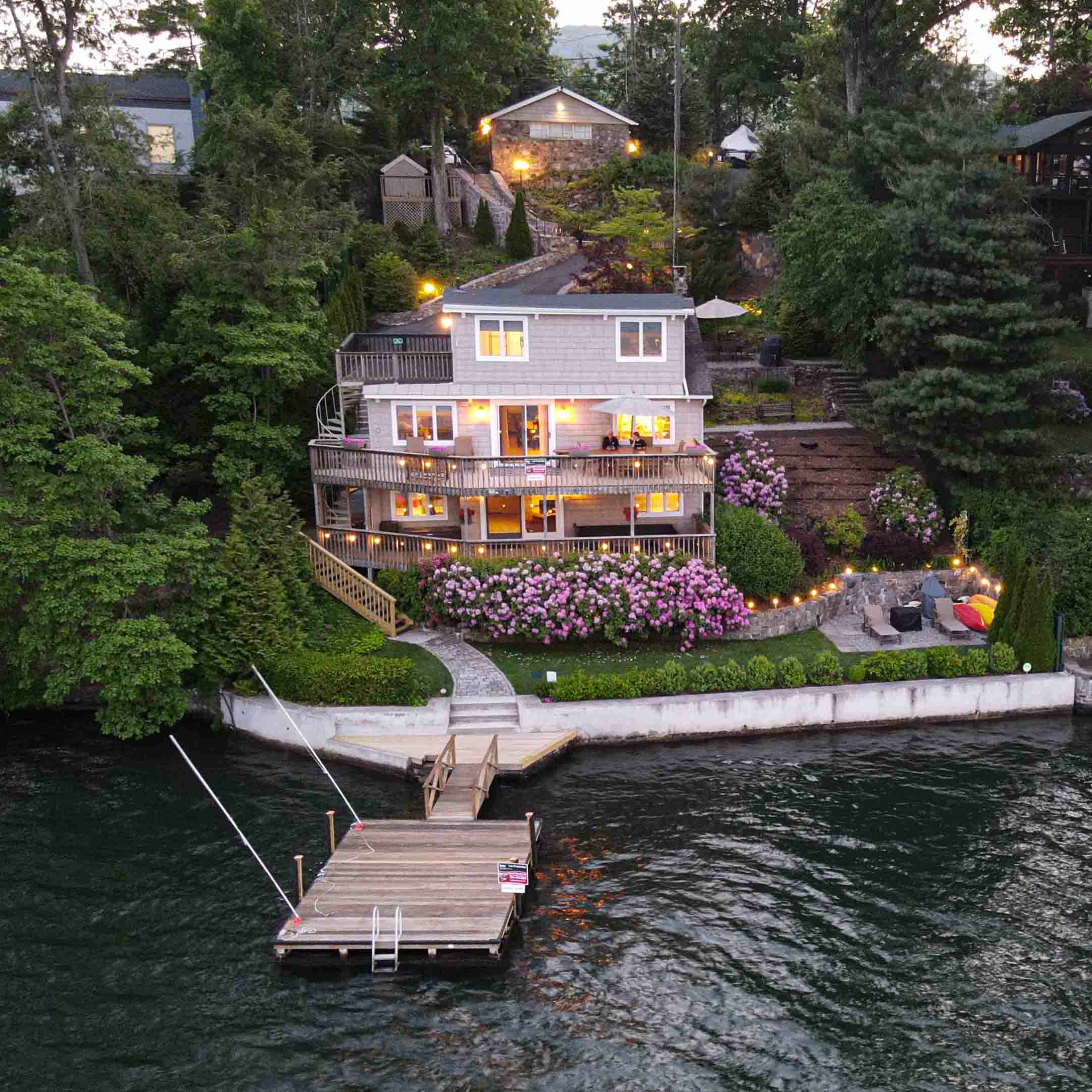 A multi-level house with large windows overlooks a lake, surrounded by lush greenery and blooming flowers, with a wooden dock extending into the water.