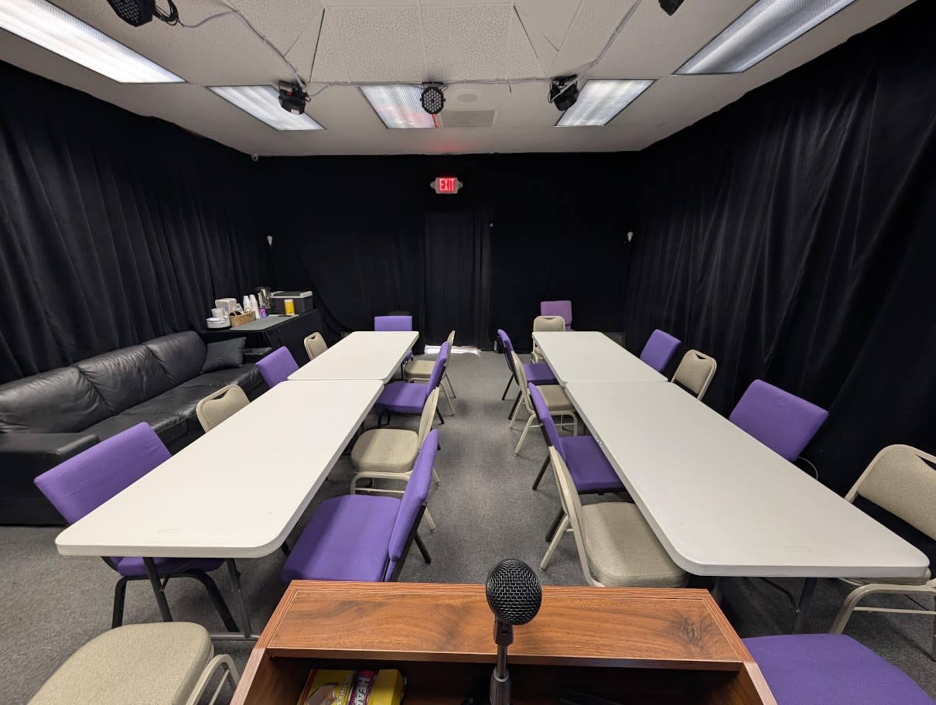 The image shows a conference room with two rows of tables arranged in a U-shape, surrounded by chairs, and a microphone stand in the foreground.