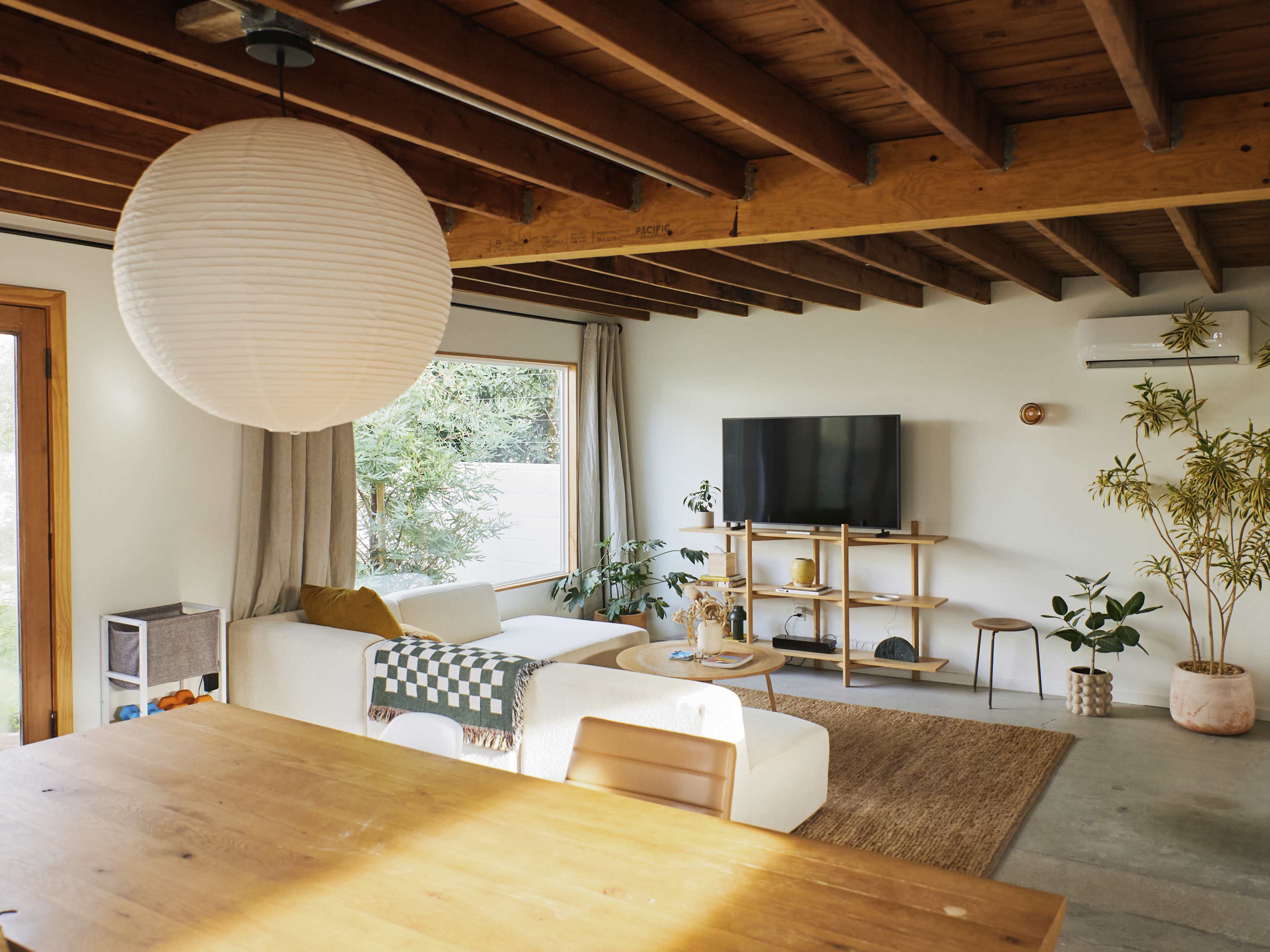 The image shows a modern living room with a light-colored sofa, a wooden coffee table, a television on a minimalist stand, and plants arranged throughout the space.