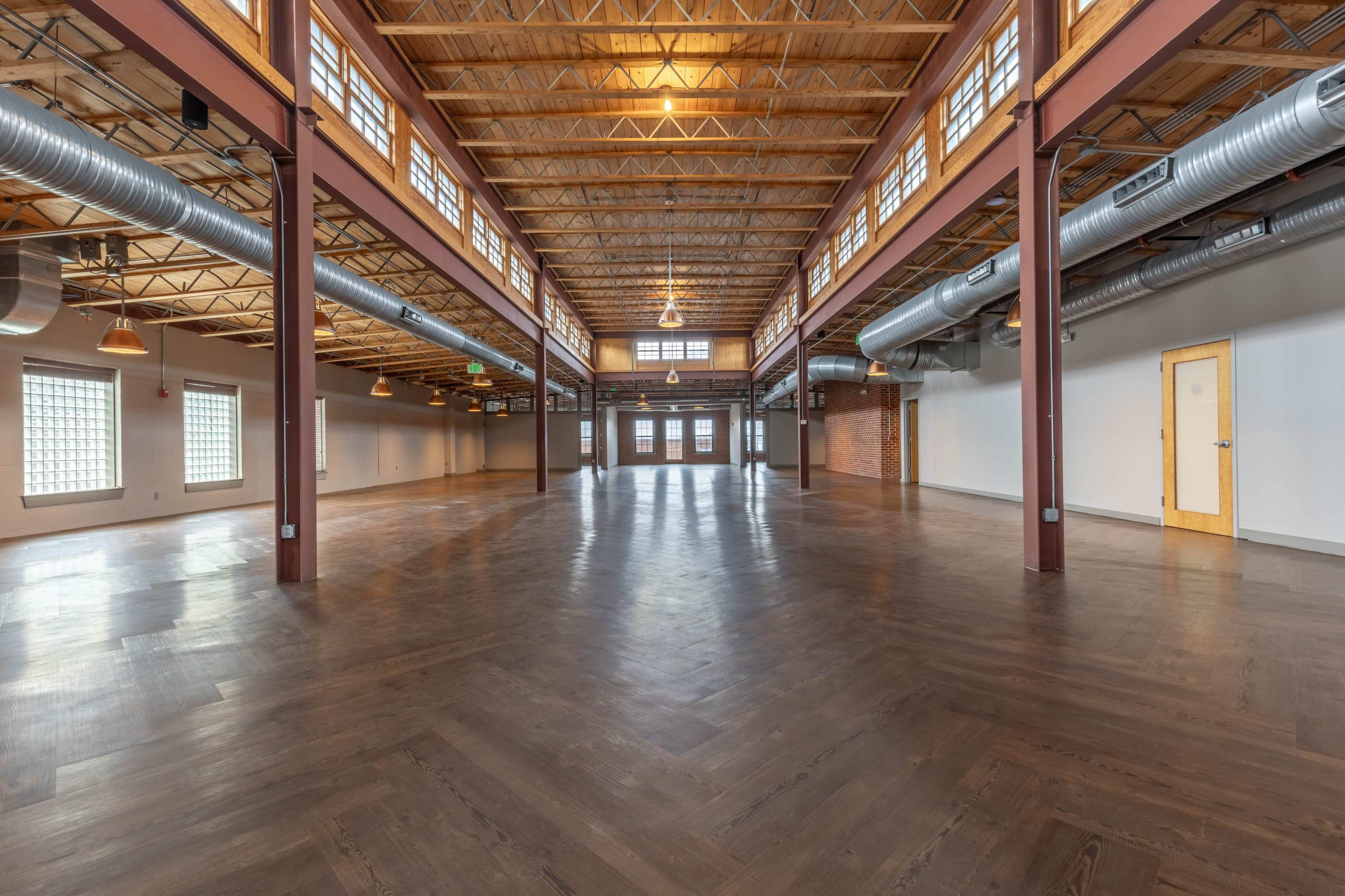 The image depicts an empty, spacious interior with wooden flooring, high ceilings, and large windows.
