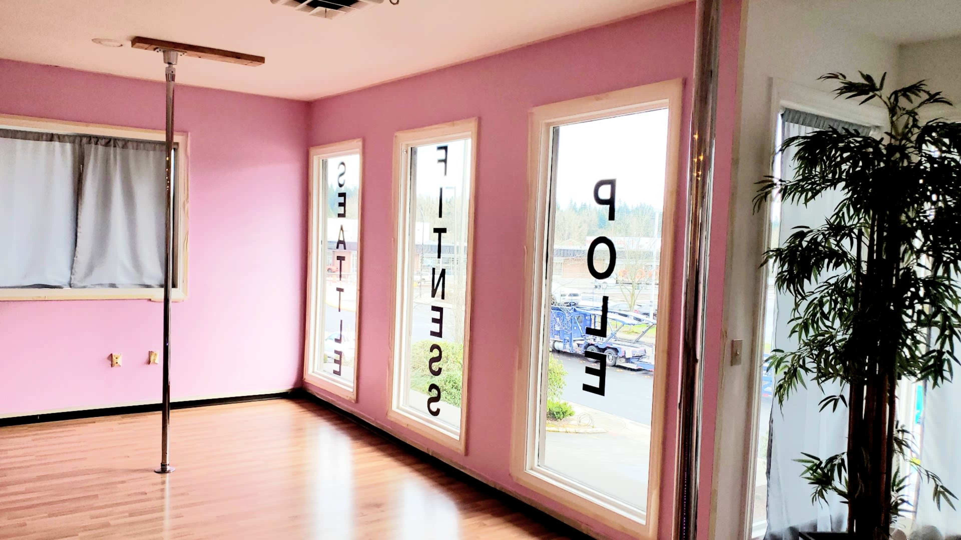 A bright room with pink walls, large windows displaying the words "FITNESS" and "STUDIO," and a pole in the center of the space.