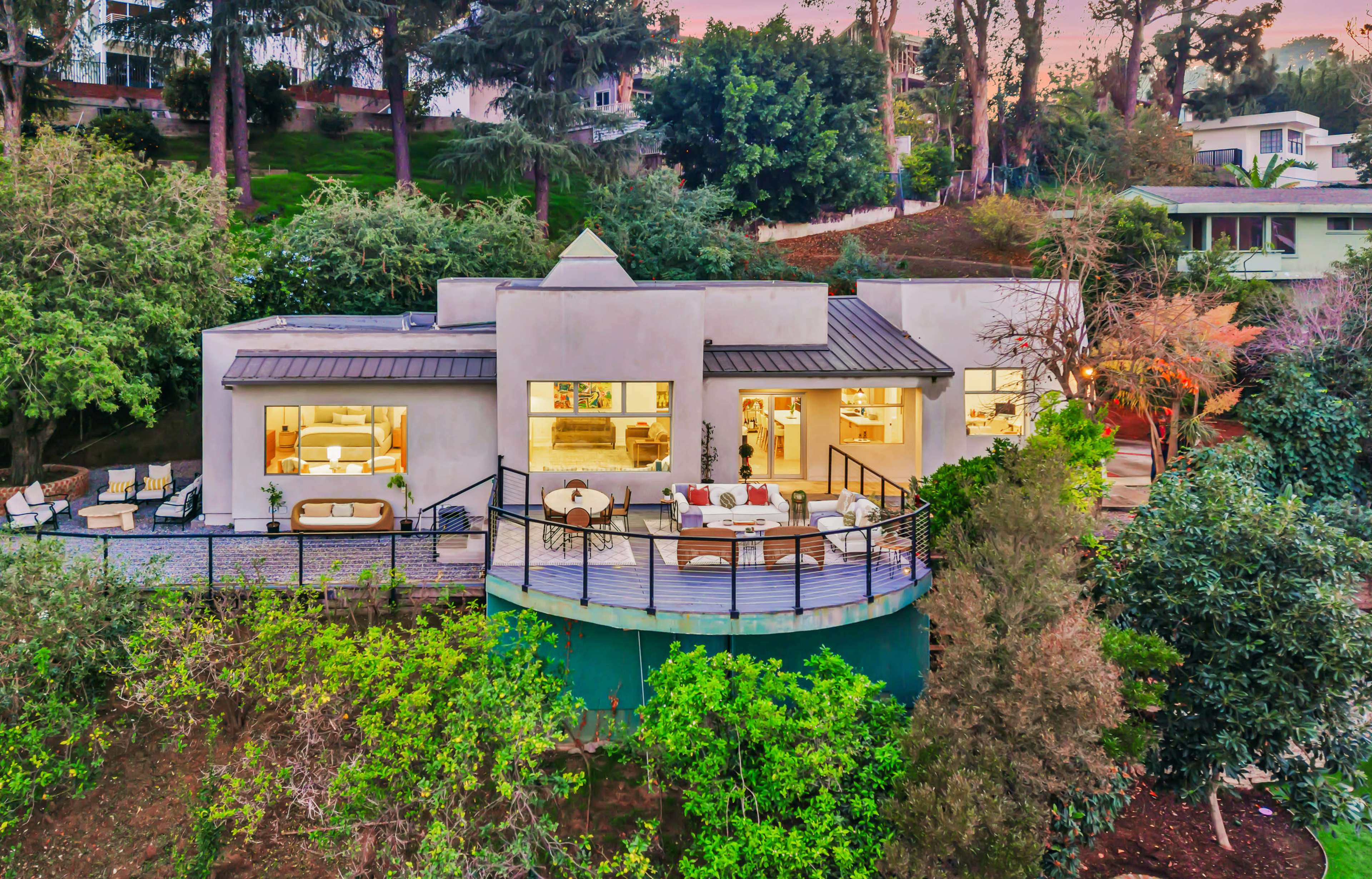 A modern house with large windows sits on a hillside surrounded by greenery and outdoor seating areas.