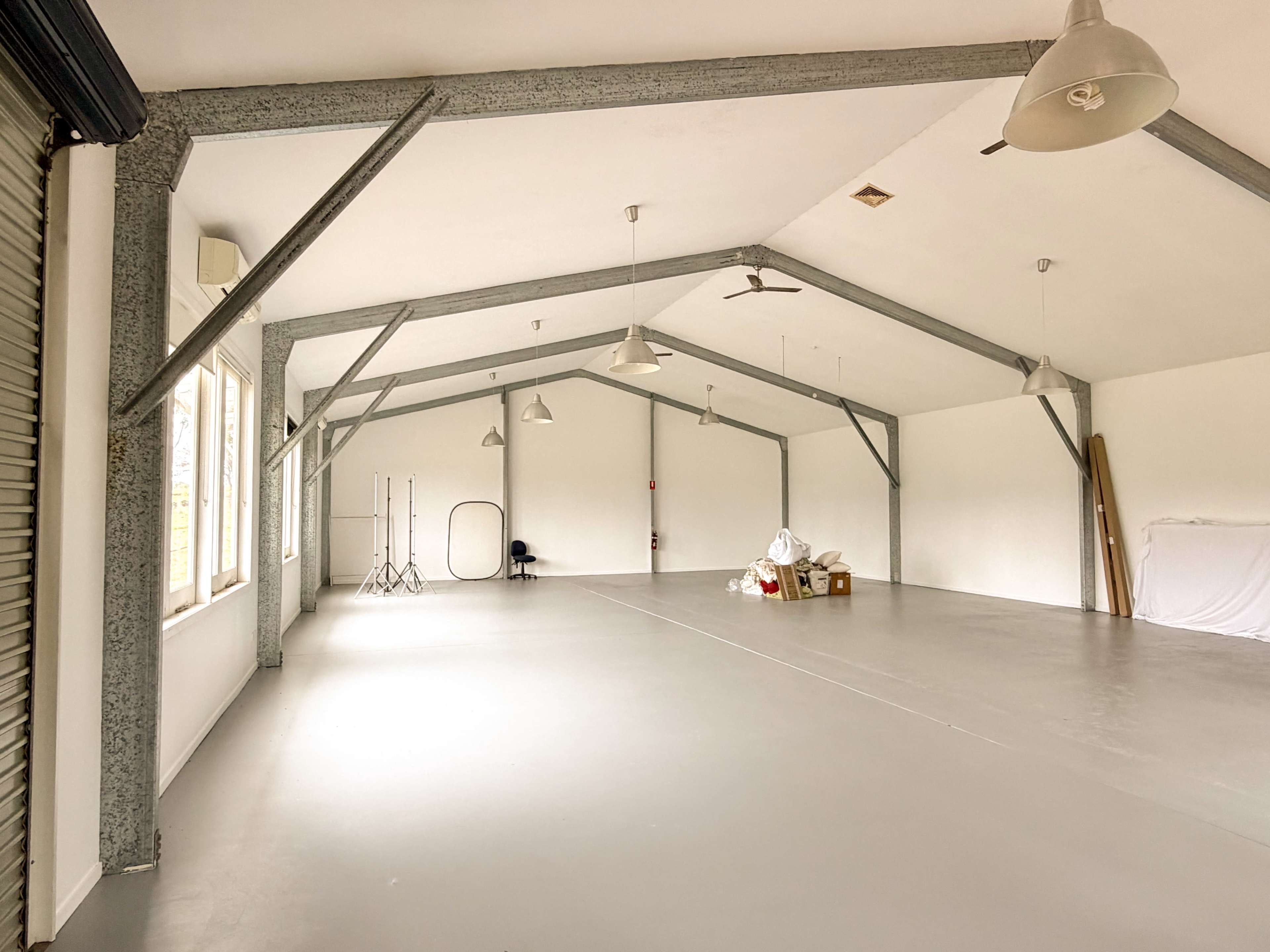 The image shows an empty, spacious room with high ceilings, metal beams, and large windows, featuring minimal furnishings.