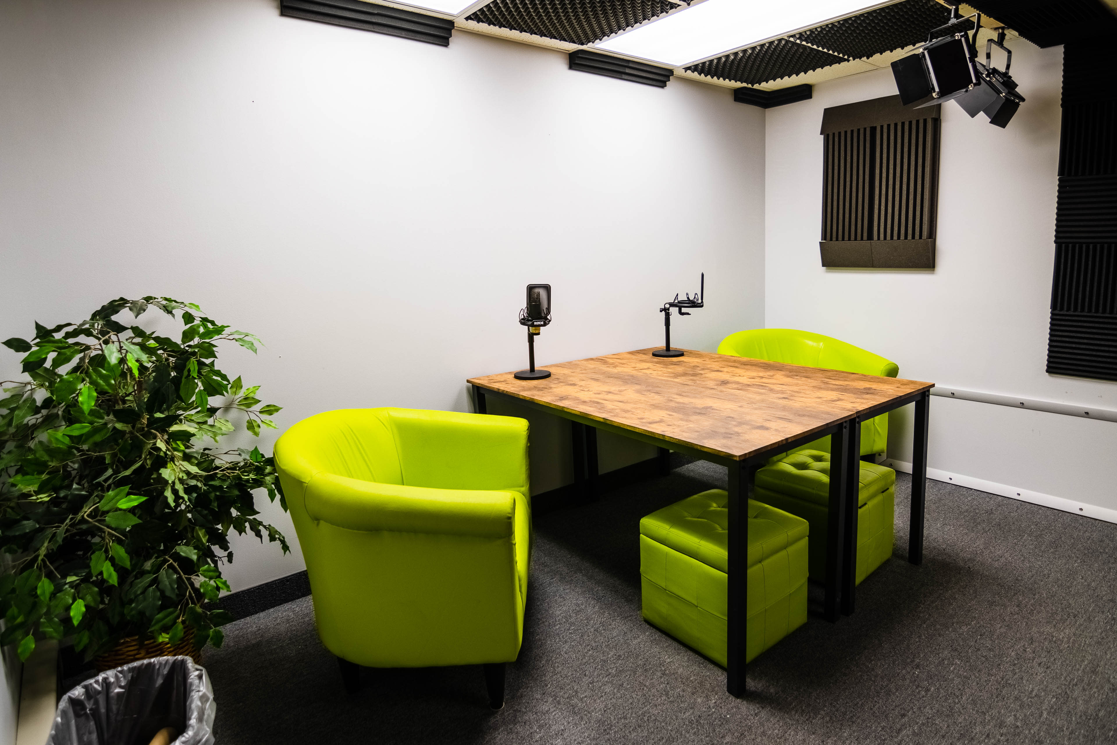 The image shows a small recording studio with a wooden table, two green chairs, microphones, and soundproofed walls.