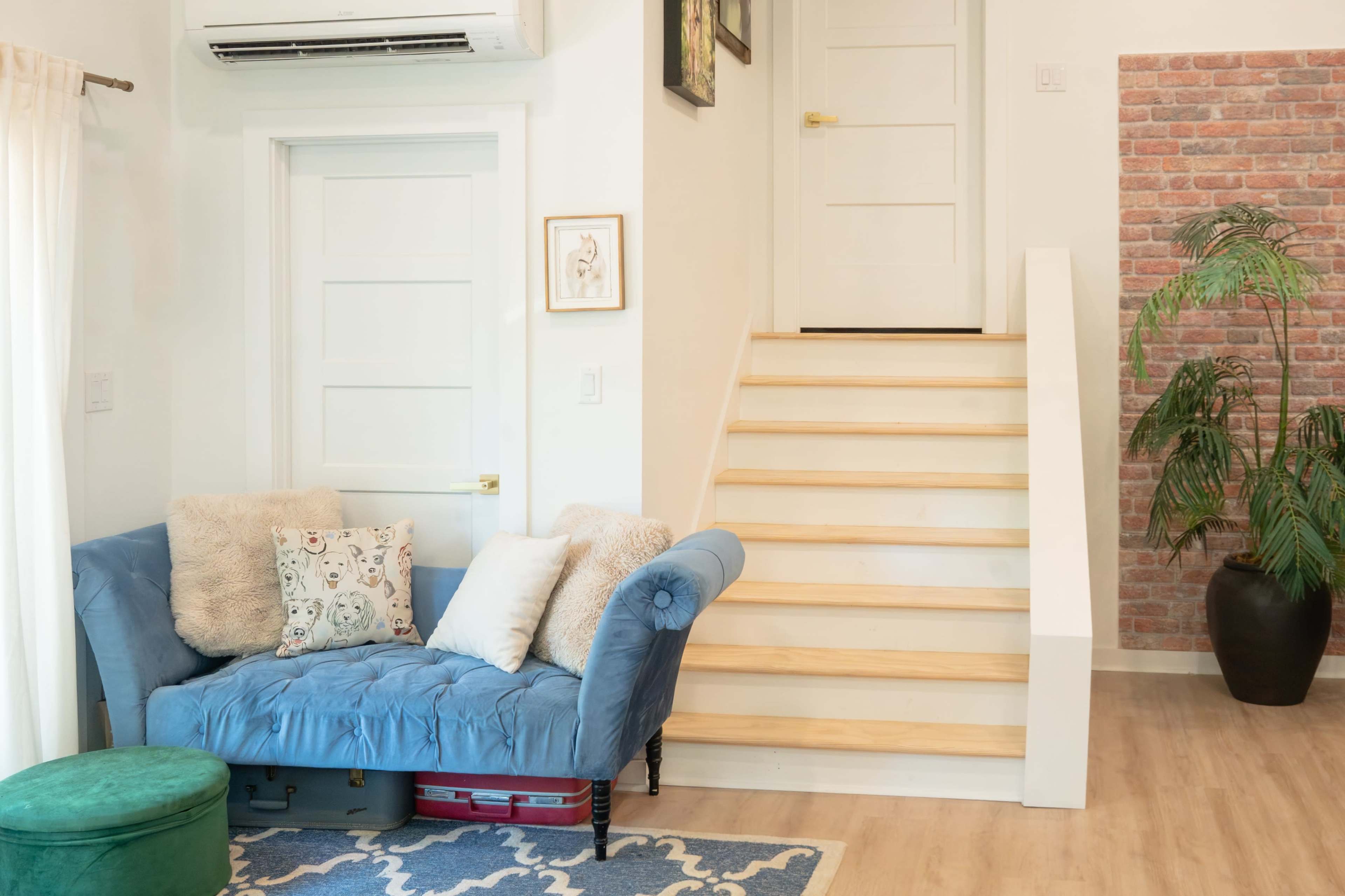 The image shows a cozy living area with a blue couch, decorative pillows, a green pouf, and a staircase leading to another level.