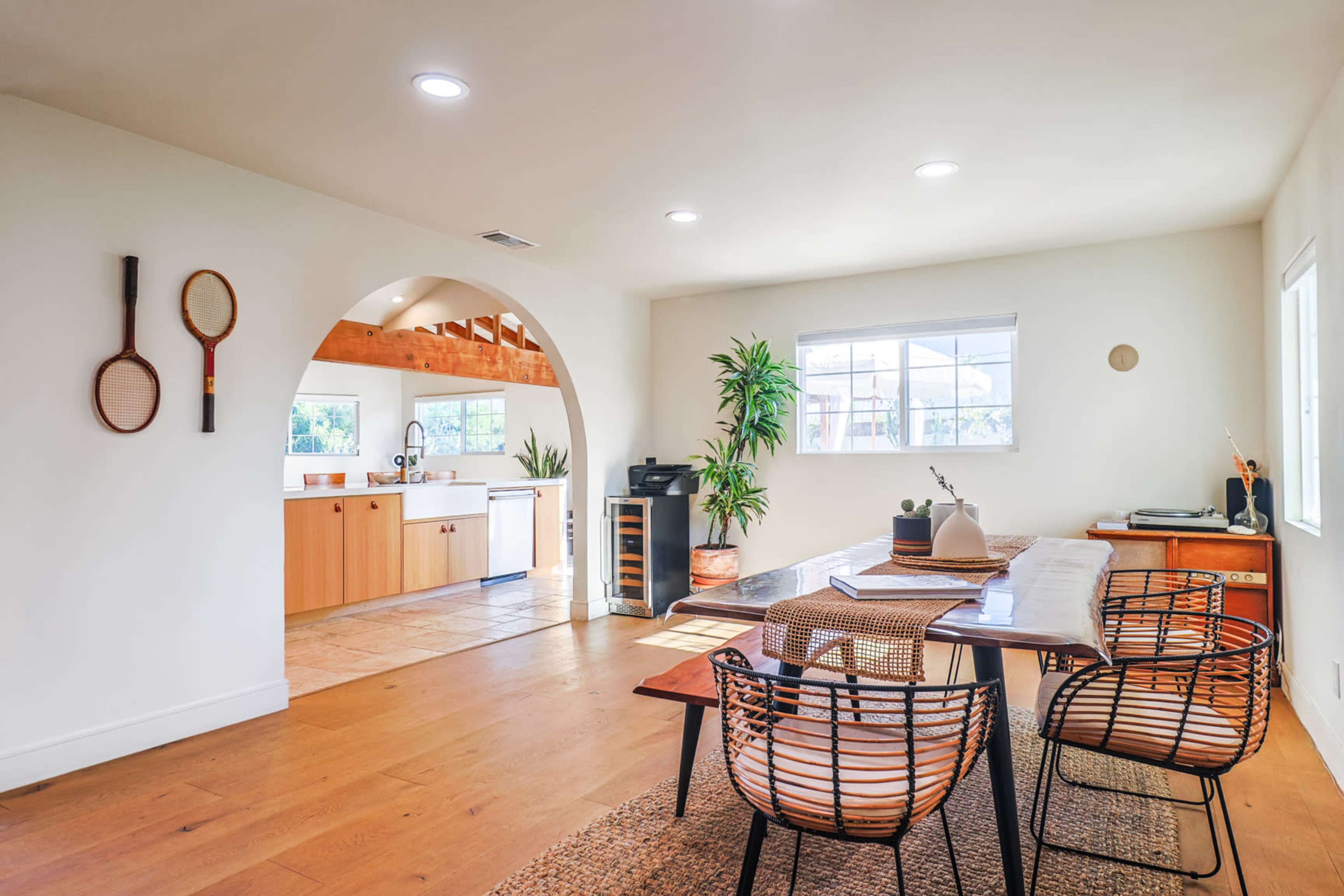 A bright, open living space featuring a wooden dining table, two woven chairs, and a kitchen visible in the background.