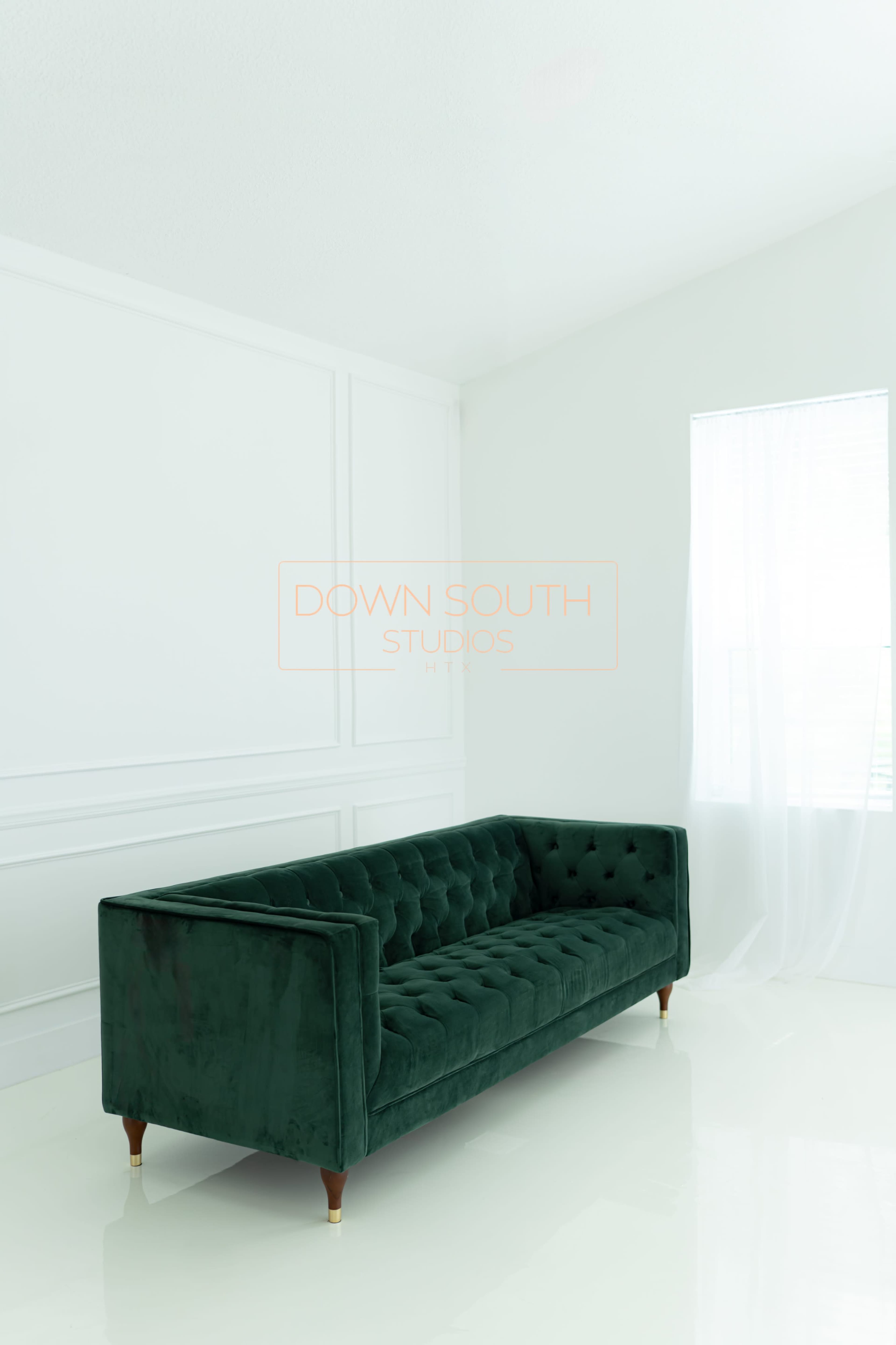 A green velvet sofa with tufted upholstery sits against a white wall in a bright, minimalistic room with a large window.