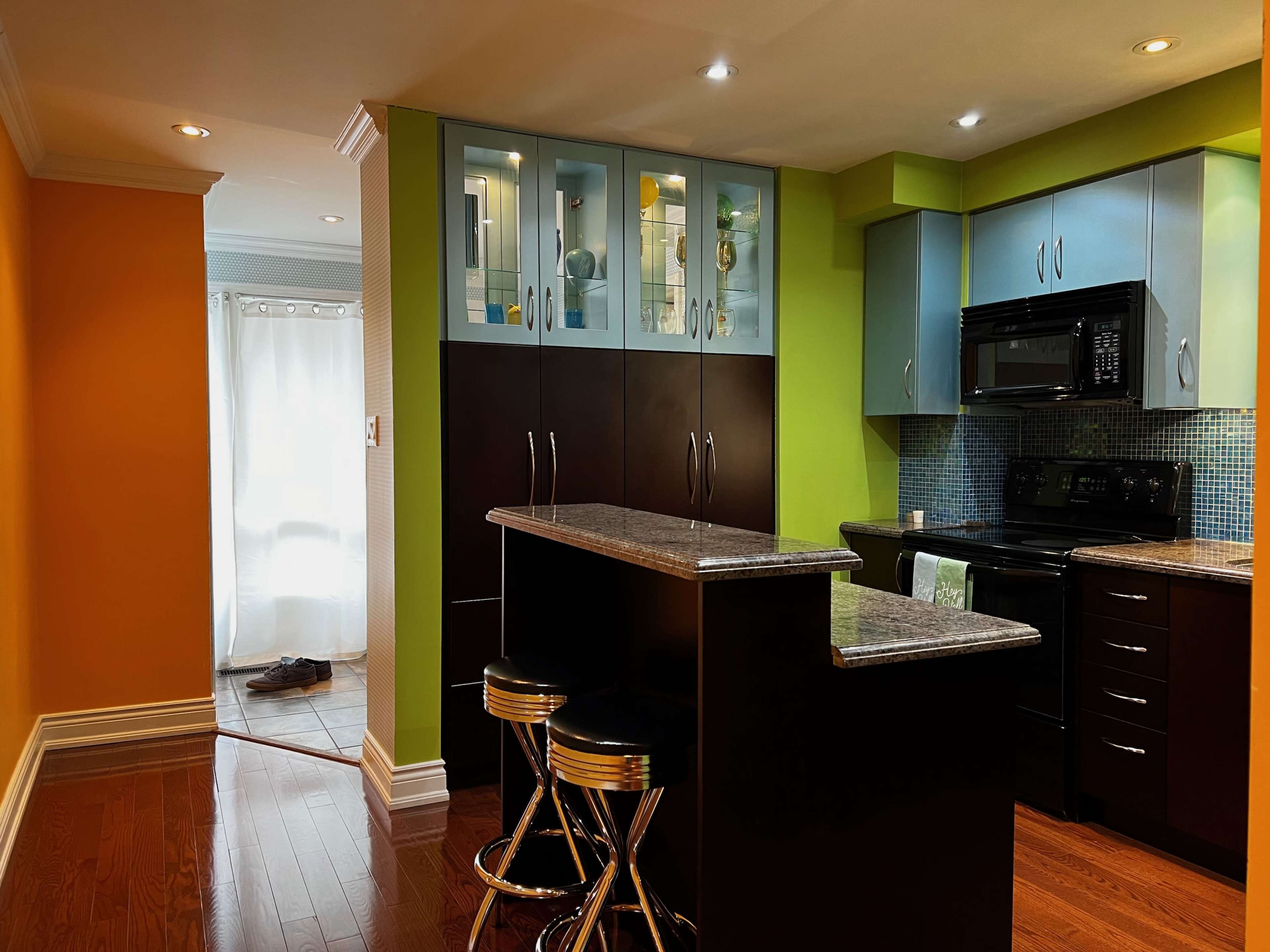 A modern kitchen area with blue and green cabinetry, a granite-topped island with two bar stools, and an adjoining brightly colored wall.