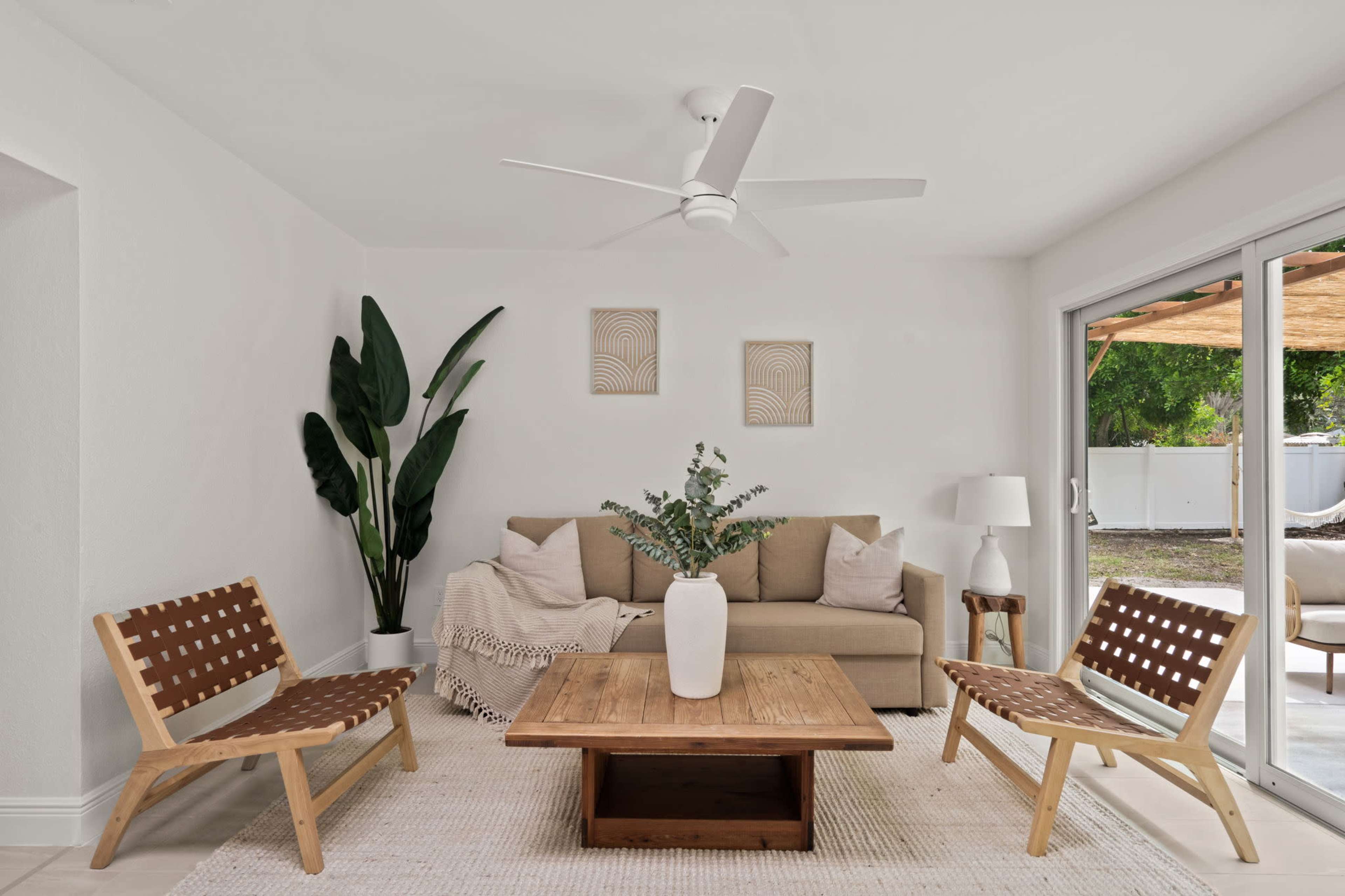 A modern living room features a beige sofa, two wooden chairs with woven seats, a wooden coffee table, and potted plants, illuminated by natural light from sliding glass doors.