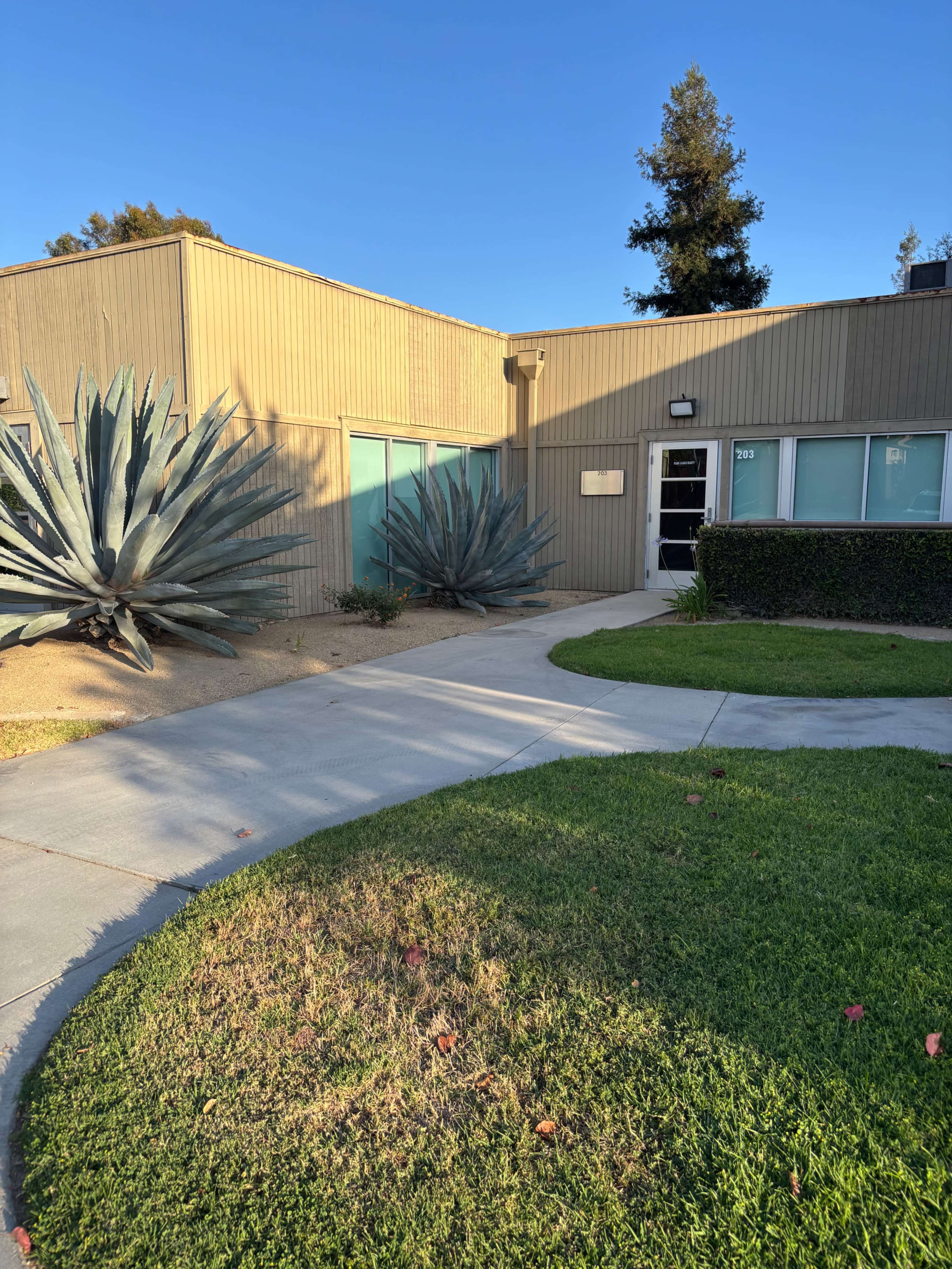 The image shows a building with large agave plants in front, alongside a curved walkway and a well-maintained lawn.