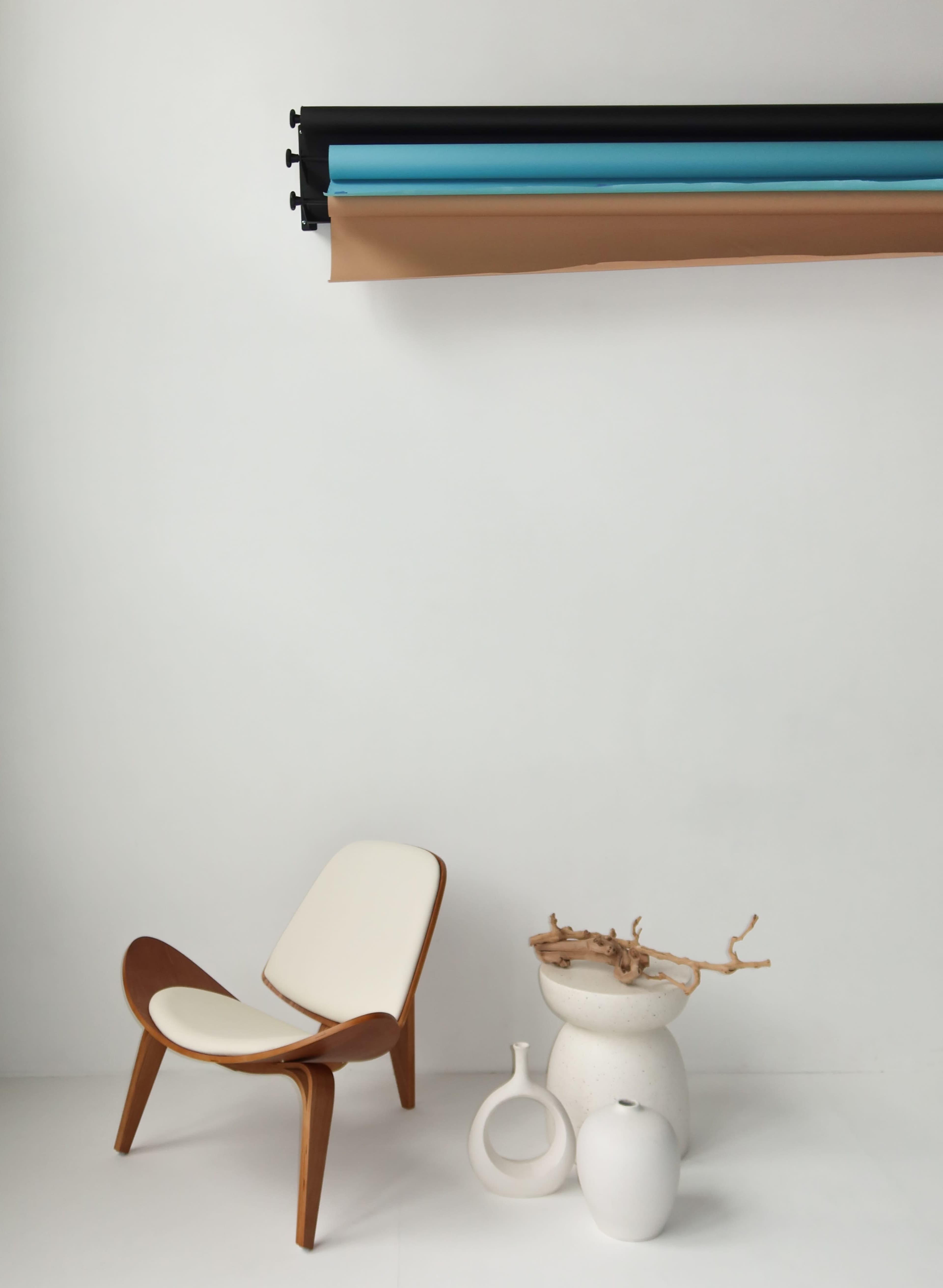 The image features a minimalist interior scene with a wooden chair, a textured white vase, and rolled paper backdrops of various colors on the wall.