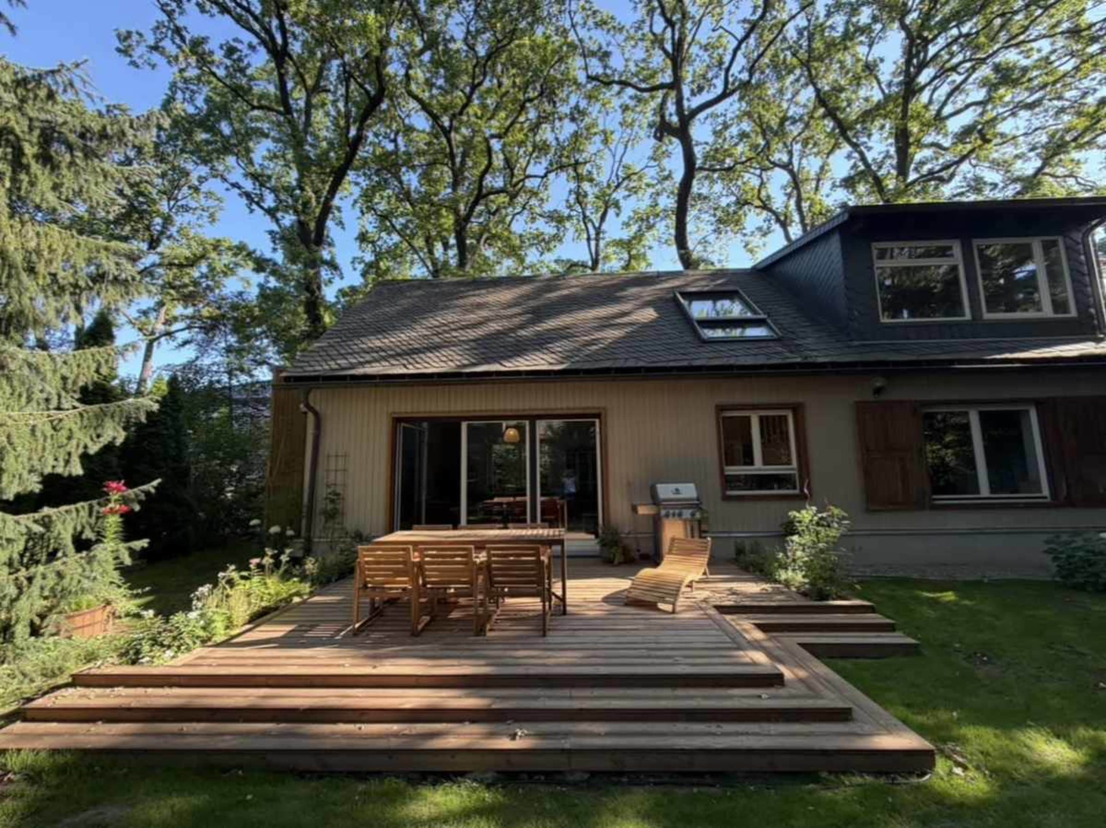 A wooden deck with a dining table and chairs is attached to a house surrounded by trees.