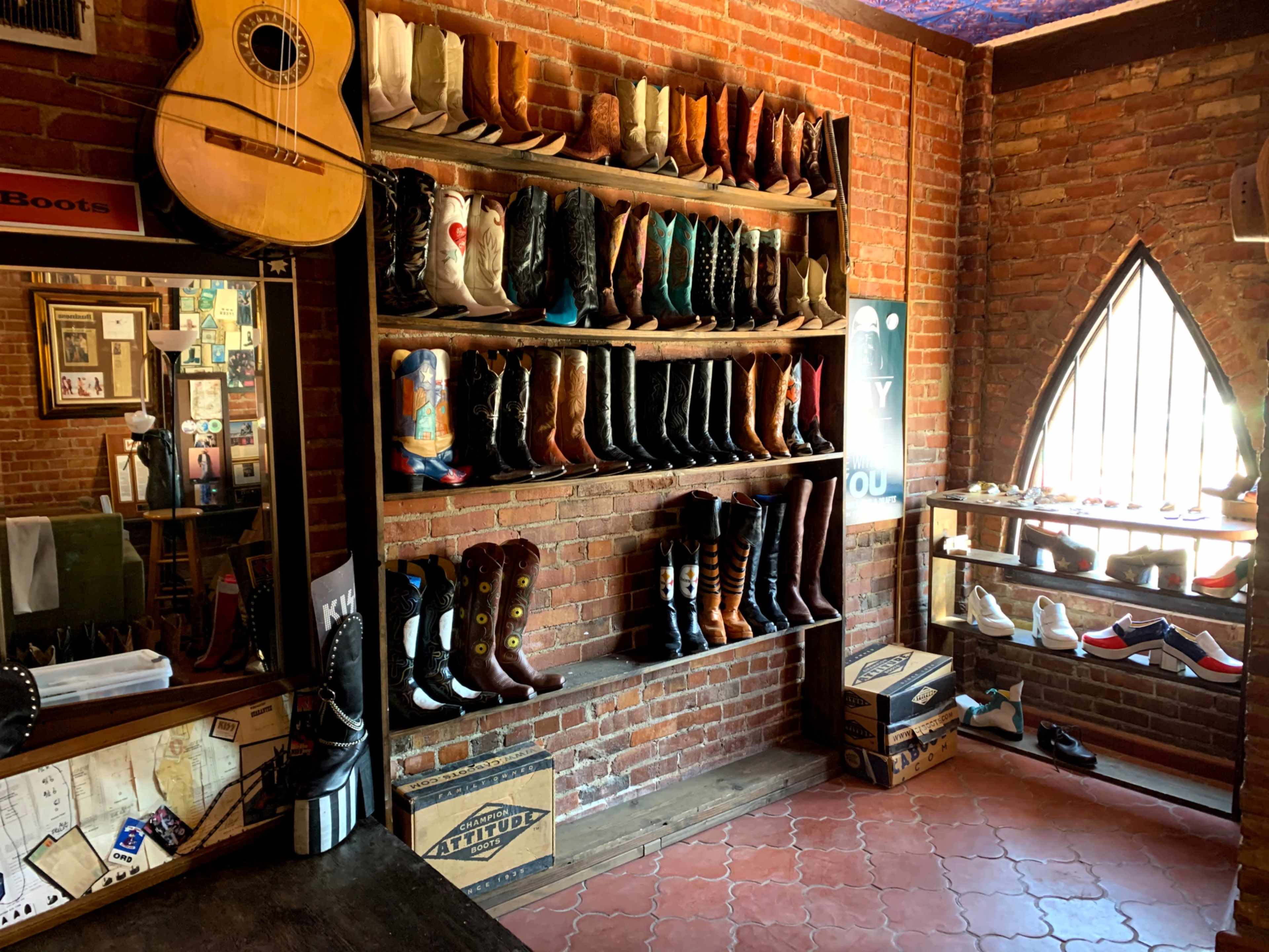 The image shows a rustic store interior featuring multiple shelves lined with various styles of boots against a backdrop of exposed brick walls.