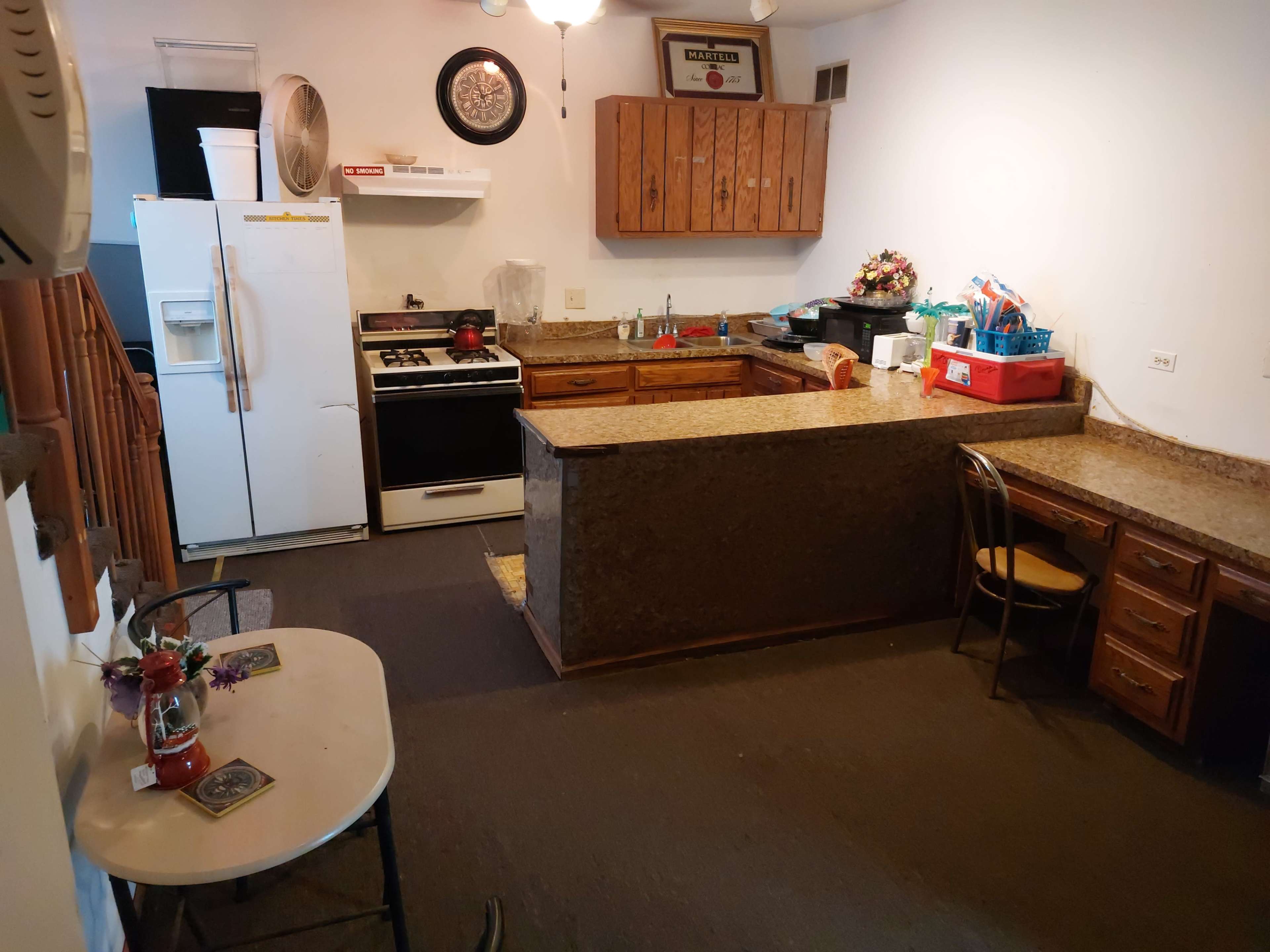 A kitchen area featuring wooden cabinets, a stove, a refrigerator, and a small dining table with a chair.