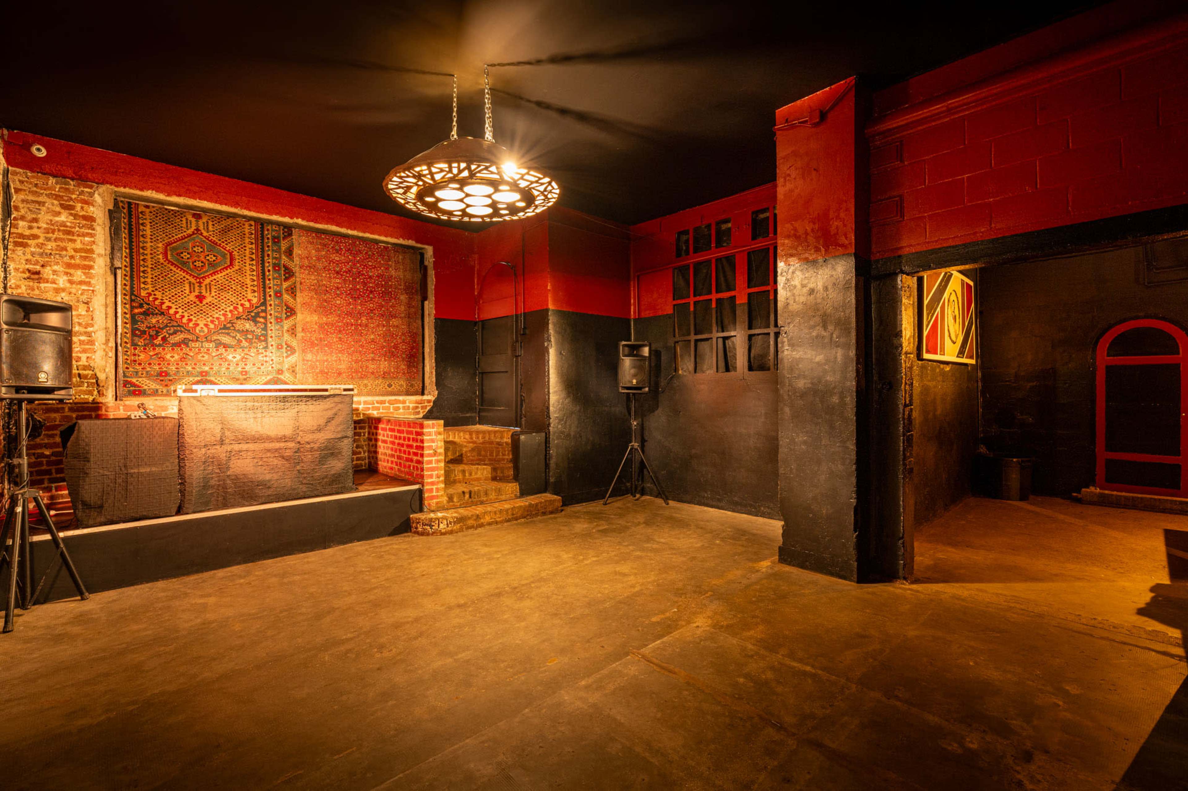An empty interior of a dimly lit venue featuring a DJ booth, red and black walls, and a staircase leading to a textured wall.